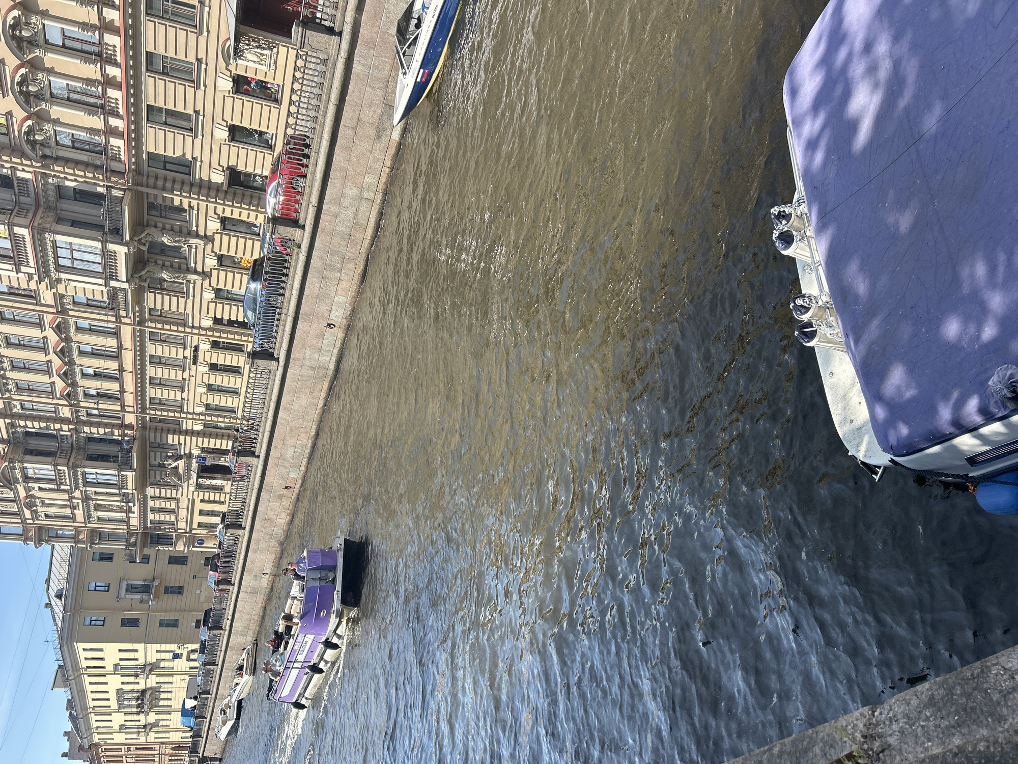 This image shows a view of a canal in a city. Several boats are visible on the water. One is a large boat with a purple covering docked on the right side of the canal, its side featuring several chrome fixtures and a blue fender. Further down the canal, a small purple boat with several people on board is traveling. Other smaller boats are also present.
On the left side of the canal is a row of multi-story buildings with ornate architecture, featuring numerous windows and decorative elements. A promenade runs along the canal, separating the buildings from the water. Cars are parked along the street behind a railing.
The water in the canal is choppy, reflecting the sunlight and the buildings. The sky appears clear and bright, suggesting it is daytime. The overall scene evokes a sense of urban life and leisure on the water. No visible text is discernible in the image.
