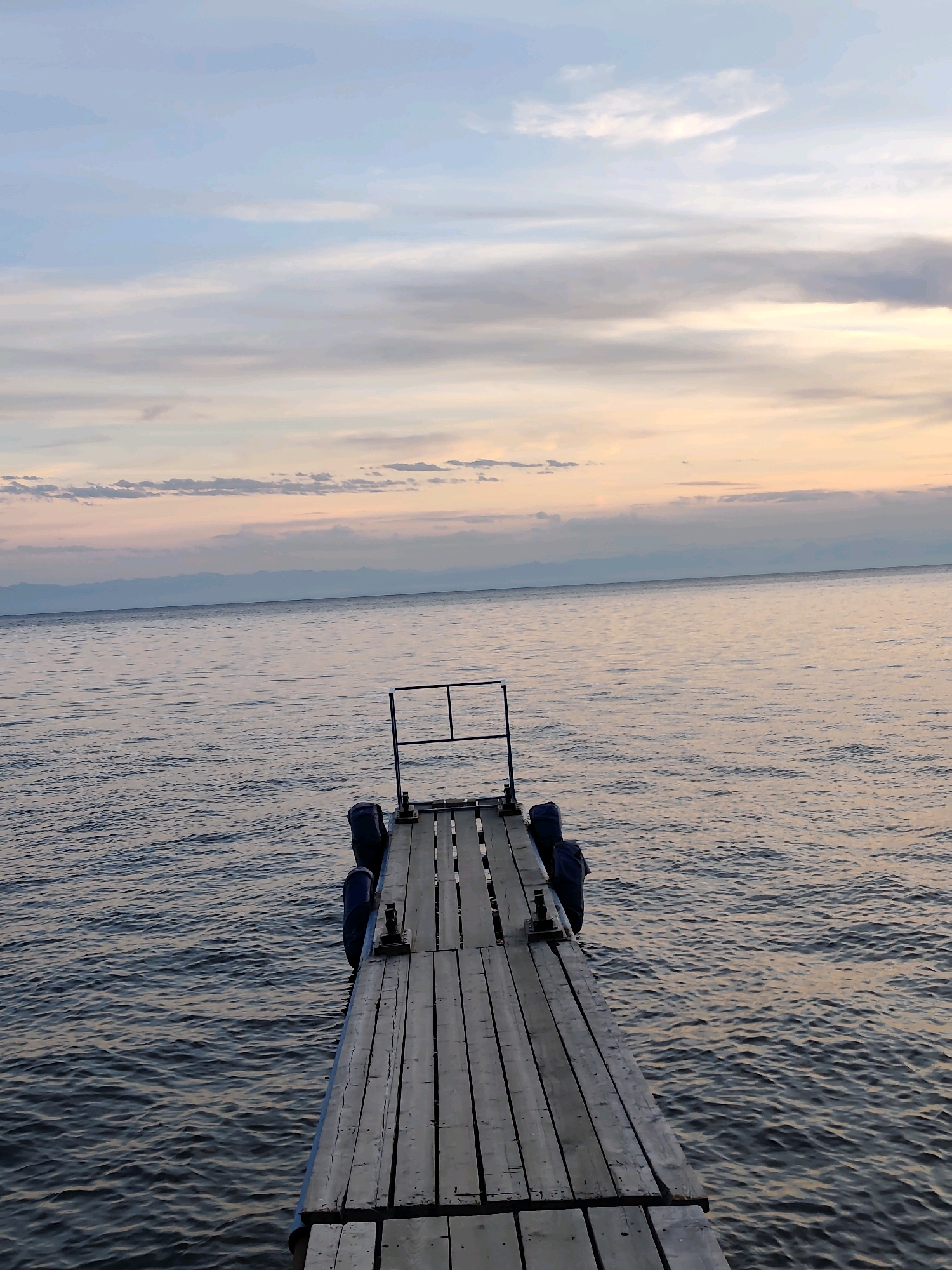 A wooden dock extends into a large body of water under a dusky sky. The dock is made of weathered planks and has a railing at its end. Blue fenders are attached to the sides of the dock. The water is calm with gentle ripples, reflecting the soft colors of the sky. In the distance, a faint outline of mountains can be seen on the horizon. The scene appears to be a tranquil evening on Lake Baikal, near Listvyanka, Russia. There are no people present in the image.