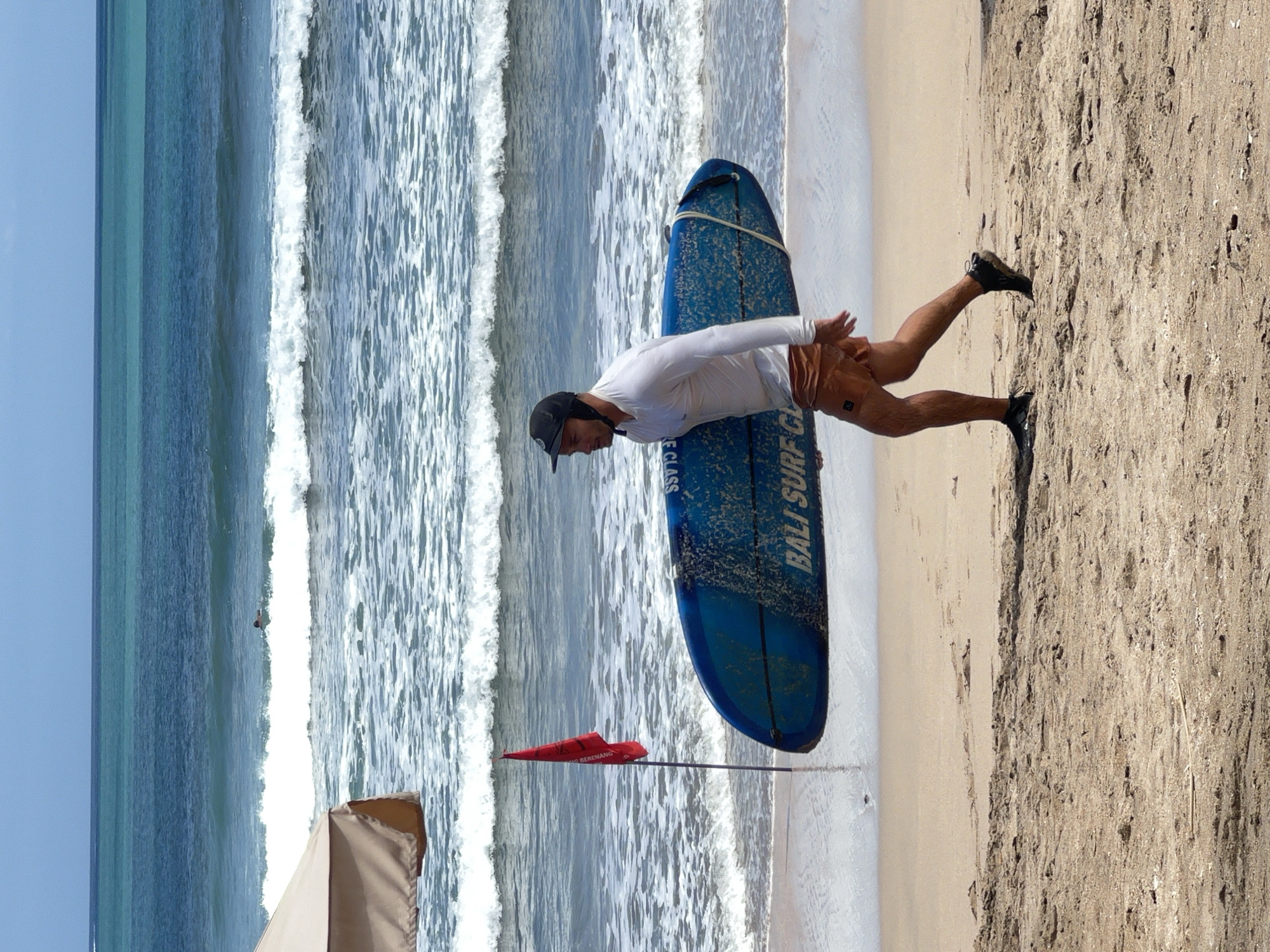 A man walks along the beach carrying a blue surfboard. He is wearing a white long-sleeved shirt and brown shorts. The surfboard has white lettering that reads "BALI SURF CLUB". A red flag is planted in the sand near the water. The ocean has waves, and there is a person surfing in the distance. The sky is blue and clear. The scene appears to be on Kuta Beach in Bali, Indonesia, a popular tourist destination known for its surf breaks and sandy shores.