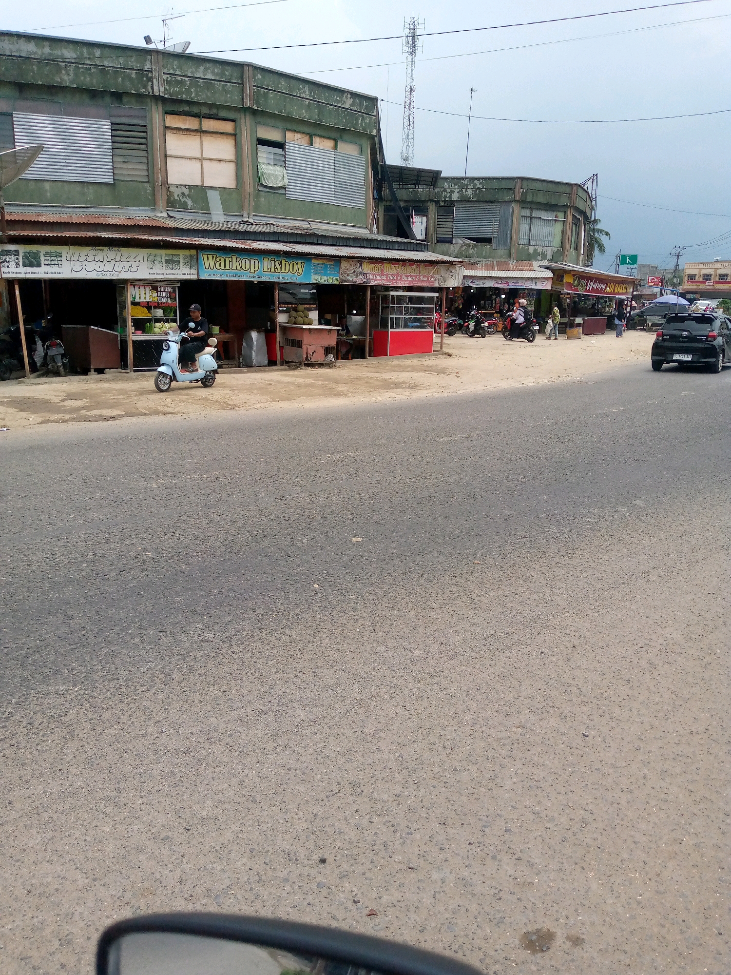 The image depicts a street scene in Keudegeudong, Indonesia, featuring a row of single-story buildings with green, weathered facades lining a dirt and asphalt road. Several commercial establishments are visible, including shops displaying signs such as "Home Made," "Warkop Lisboy," and "Warung Adi Bakso." Various goods are displayed outside the shops, including fruits like durian, and there are several motorcycles parked. A light blue scooter is in the foreground with a person wearing a black t-shirt and cap seated on it, facing left. A black sedan is driving away on the right side of the road. Overhead, power lines and a telecommunications tower are present against a cloudy sky. The environment appears to be a typical roadside commercial area with evidence of both vehicular and pedestrian activity.