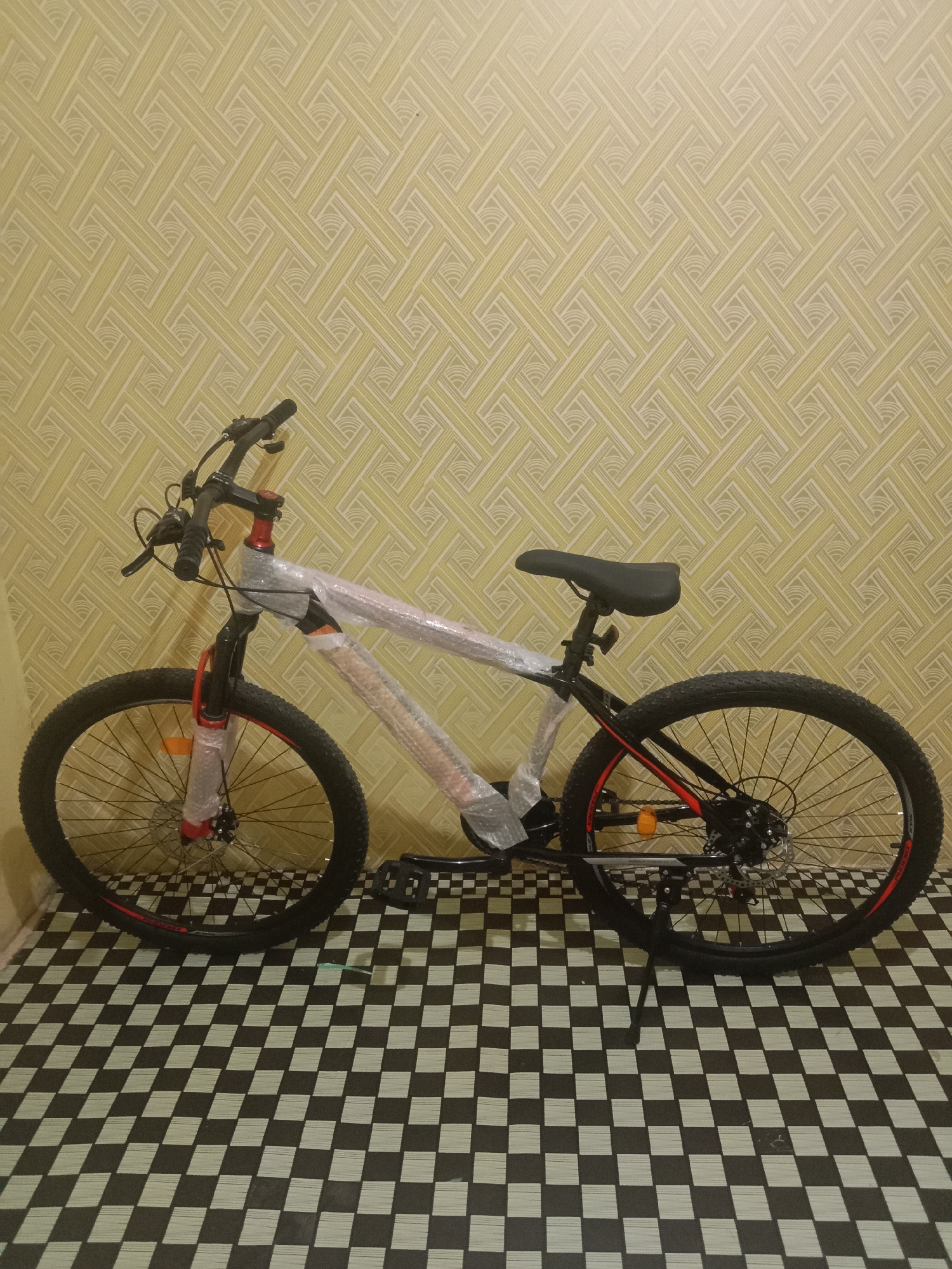 An unassembled bicycle is propped up on its kickstand against a patterned yellow wall. The bicycle is wrapped in clear plastic protective covering, indicating it is new or being transported. The handlebars are turned to the left, and the seat is a standard black bicycle saddle. The front wheel is on the left side of the image, and the rear wheel is on the right, showing a disc brake system. The floor is a black and white checkered pattern. The overall scene suggests a domestic indoor setting, potentially a home in Kutosari, Indonesia, where the bicycle is being stored or prepared.
