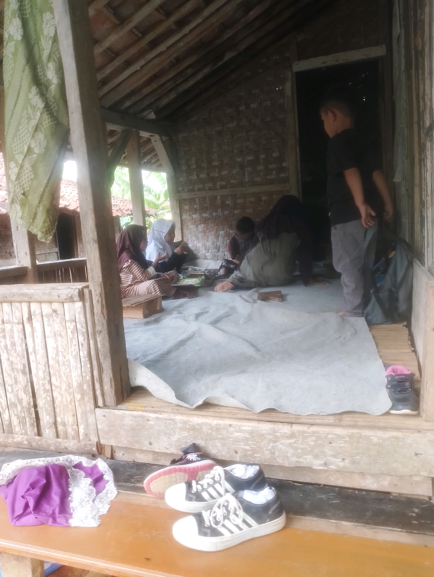 In Kalapadua, Indonesia, an open-air structure with a thatched roof and bamboo walls serves as a gathering space. Inside, four individuals are present: two young females in hijabs, one wearing a brown and white striped outfit and the other a white hijab and black garment, appear to be reading from books placed on small wooden stands. Another individual, a young boy in a black t-shirt and grey pants, stands near a doorway. A fourth person, whose gender is unclear and who is wearing dark clothing, is seen seated on the floor in the background, possibly engaged in reading or prayer. A large grey sheet covers most of the floor, with a blue backpack situated to the right of the boy. In the foreground, resting on a wooden table, are a purple and white fabric item and two pairs of sneakers, one black and white with white laces, and another partially visible black shoe with pink accents. The overall scene suggests an informal educational or communal activity taking place within a rustic, open-air setting.