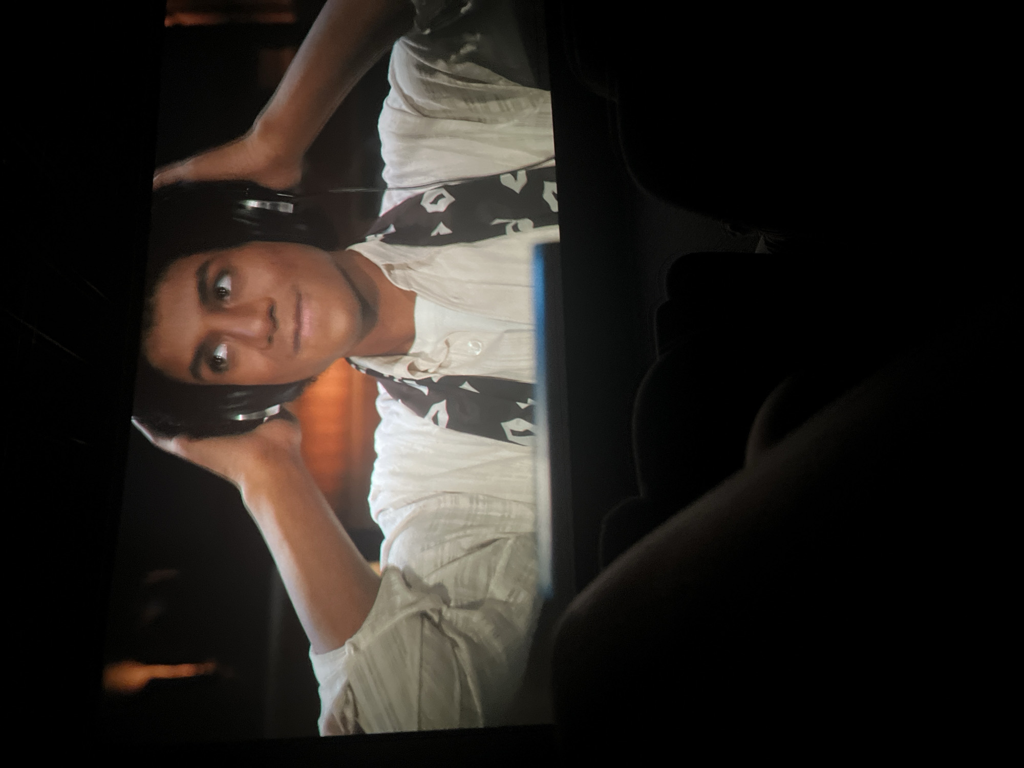 In a dimly lit, indoor environment resembling a movie theater, a young individual with dark hair is captured on a screen, wearing headphones and a light-colored, long-sleeved shirt with a patterned scarf. The individual's gaze is directed upwards and to the left. The foreground of the image is dominated by the dark silhouettes of what appear to be people seated in front of the screen, with a portion of one person's arm and hand visible. The screen itself is positioned low, with a dark border at the top and bottom.