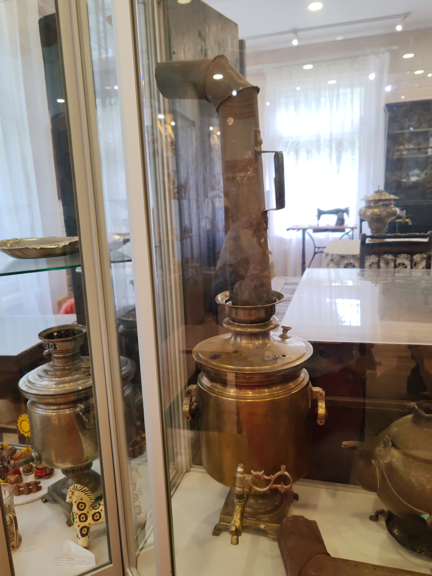 The image depicts an interior scene, likely a museum exhibit, featuring several antique samovars displayed behind glass. The central focus is a large, ornate brass samovar with a prominent pipe extending upwards. To the left, another silver-colored samovar is partially visible within a display case, alongside decorative items. In the background, glimpses of a sewing machine and other display items can be seen. The overall environment suggests a cultural or historical exhibition space. The photograph was taken in Vizinga, Russia.
