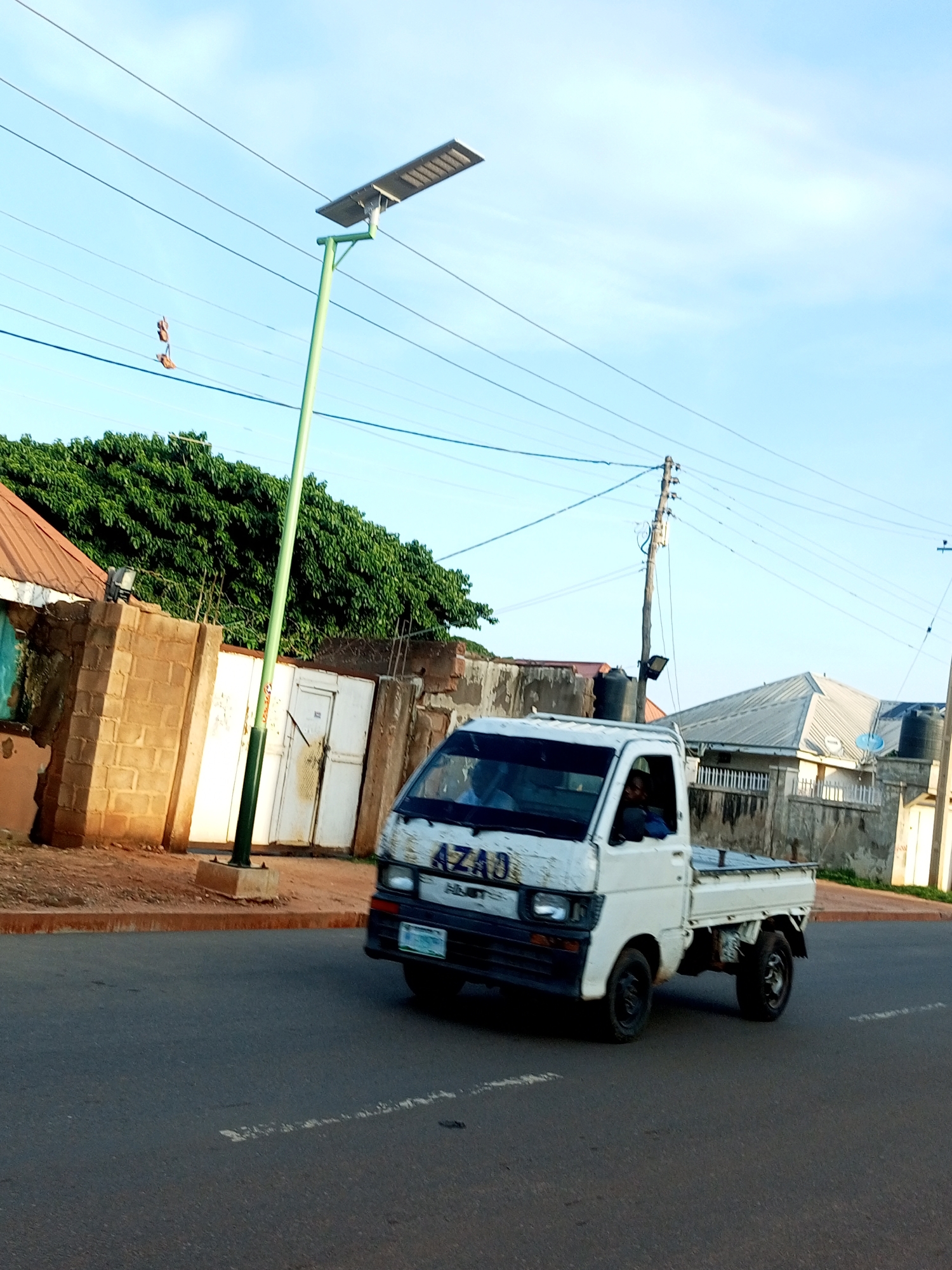 In Tempure, Nigeria, an image captures a view of a street with a white truck driving by. The truck has "AZAD" written on its side. To the left of the truck, there's a solar-powered street light and a brick wall with a gate. In the background, a dense green tree stands tall, and further in the distance, several buildings with corrugated roofs are visible. Power lines crisscross the sky, and a utility pole with a transformer is also present. The sky is a clear blue with a few wispy clouds.