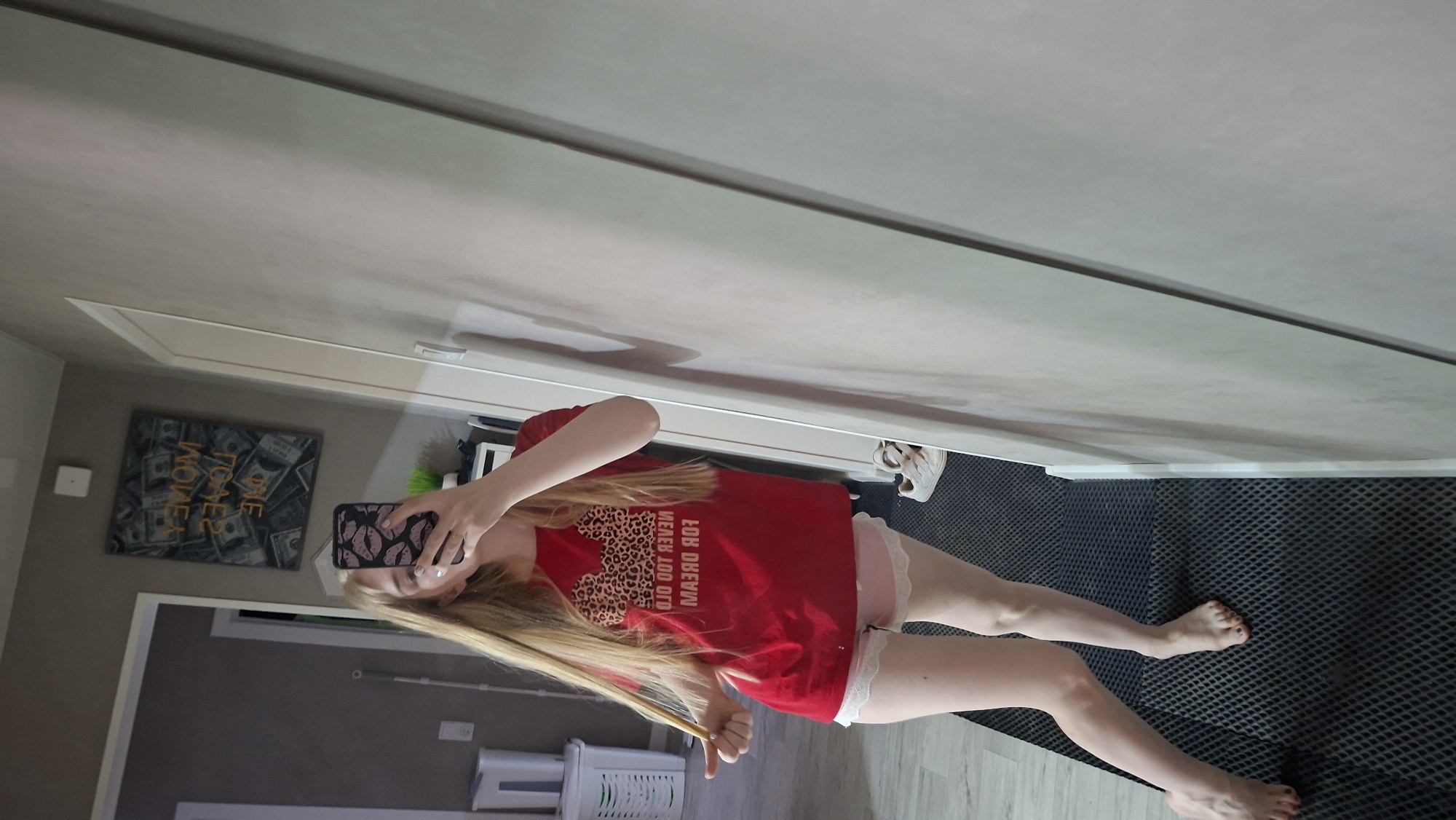 A mirrored perspective shows a young woman with long blonde hair and a red t-shirt with leopard print on the chest and text that appears to be mirrored. She is holding up a phone with a pink patterned case, obscuring her face. She is wearing pink shorts with white lace trim. She is barefoot, standing on a grey mat. Behind her is a hallway with a picture of money on the wall. A white laundry hamper is visible in the background. The location is Chaikovsky, Russia.