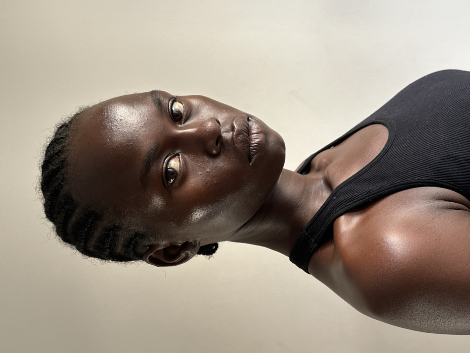 A close-up portrait features a female individual with dark skin, dark eyes, and dark hair styled in cornrows. She is positioned facing forward, slightly angled to her right, and gazes directly at the viewer with a neutral expression. Her visible attire consists of a black, ribbed tank top or camisole. The subject's skin exhibits a distinct sheen, particularly on her forehead, cheekbones, and the exposed areas of her shoulders and clavicle. The background is a plain, uniformly lit, light-colored wall, devoid of any discernible objects, patterns, or contextual elements that would indicate a specific location in Paris, France, or any other city.