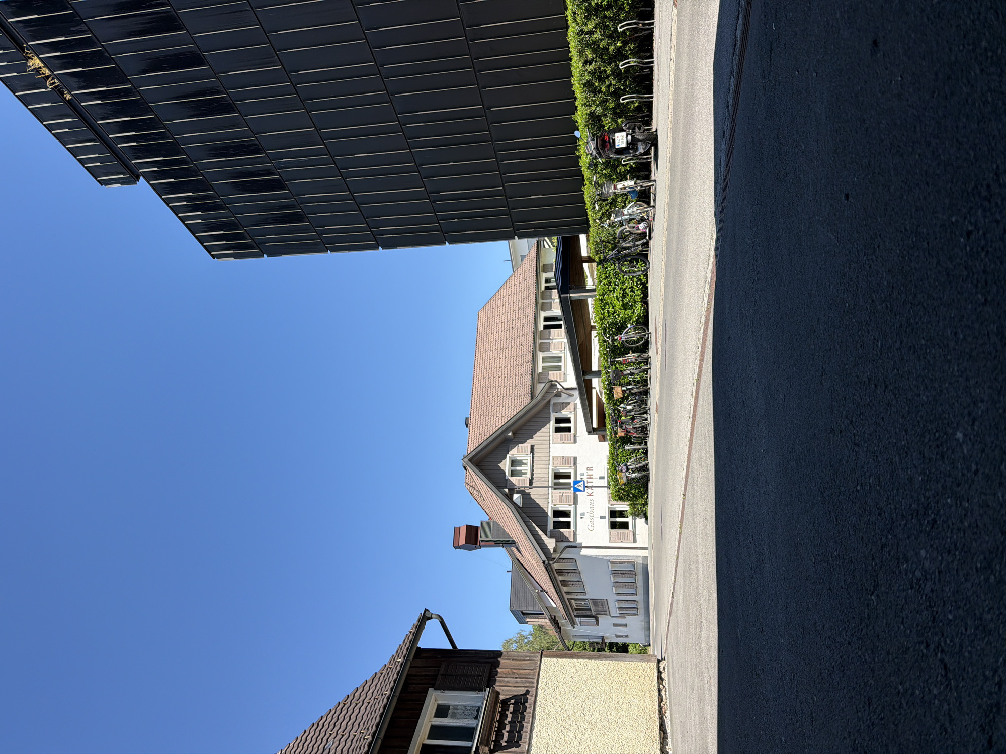 The image captures a street scene in the city of Hard, Austria, on a clear, sunny day. In the foreground, a dark asphalt road takes up the lower portion of the frame, with a distinct shadow cast from an unseen object on the left. To the left of the road, a building with a light yellow textured facade and a brown tiled roof is partially visible. Adjacent to this, a white building with brown shutters and a red-tiled roof is situated. A sign on this building reads "Gasthaus KATH'R". In front of the Gasthaus KATH'R, a line of bicycles and a scooter are parked along a low, dense green hedge. To the right of the Gasthaus KATH'R, a modern, tall black building with vertical paneling dominates the right side of the image, reaching towards the bright blue sky. The overall environment suggests a commercial or residential area with a mix of traditional and contemporary architecture.