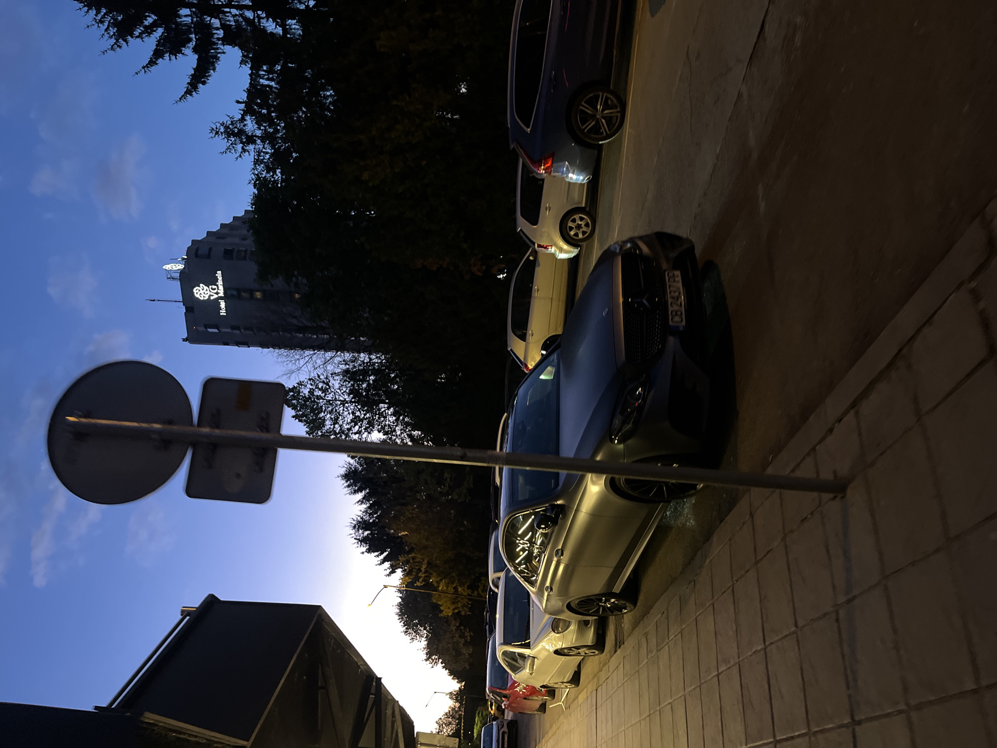 The photograph, captured at dusk or twilight, presents an urban street scene with a tall hotel building and parked cars. The image appears to be rotated 90 degrees counter-clockwise, showing a deep blue sky with faint clouds.
On the left side of the frame, a prominent, multi-story building stands against the twilight sky. Its illuminated signage clearly displays "Hotel Marinela" with a distinctive "VG" logo above the text. Silhouetted trees frame parts of the building and the sky, indicating lush foliage.
Along the right side of the image, a row of cars is parked on a paved surface, likely a sidewalk or parking area, which features rectangular block patterns. The closest car is a dark-colored sedan, and its front license plate is visible, reading "CB 2437 HP." Behind it, a silver SUV/wagon, a white SUV, and other vehicles are parked in sequence.
In the mid-ground, a metal pole supports the rear of a circular sign and a rectangular sign, silhouetted against the sky. The scene is devoid of people and depicts no specific activity, conveying a calm and still moment in an urban environment. The license plate "CB 2437 HP" confirms the location context of Sofia, Bulgaria.