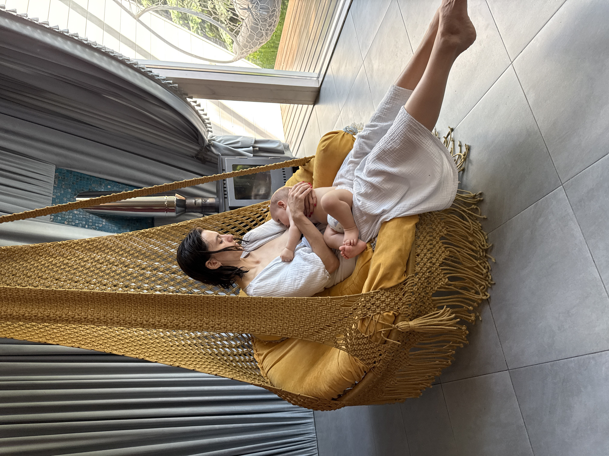 A woman lies in a hammock, holding a baby. The hammock is made of a golden rope material with yellow cushions. The woman is wearing a white robe. The baby is wearing a diaper. The room has gray tiled floors and gray curtains. There is a window behind the hammock, and what appears to be a heater. The location is a spa. There is no definitive proof of the location in the city of Чёрное, Russia.