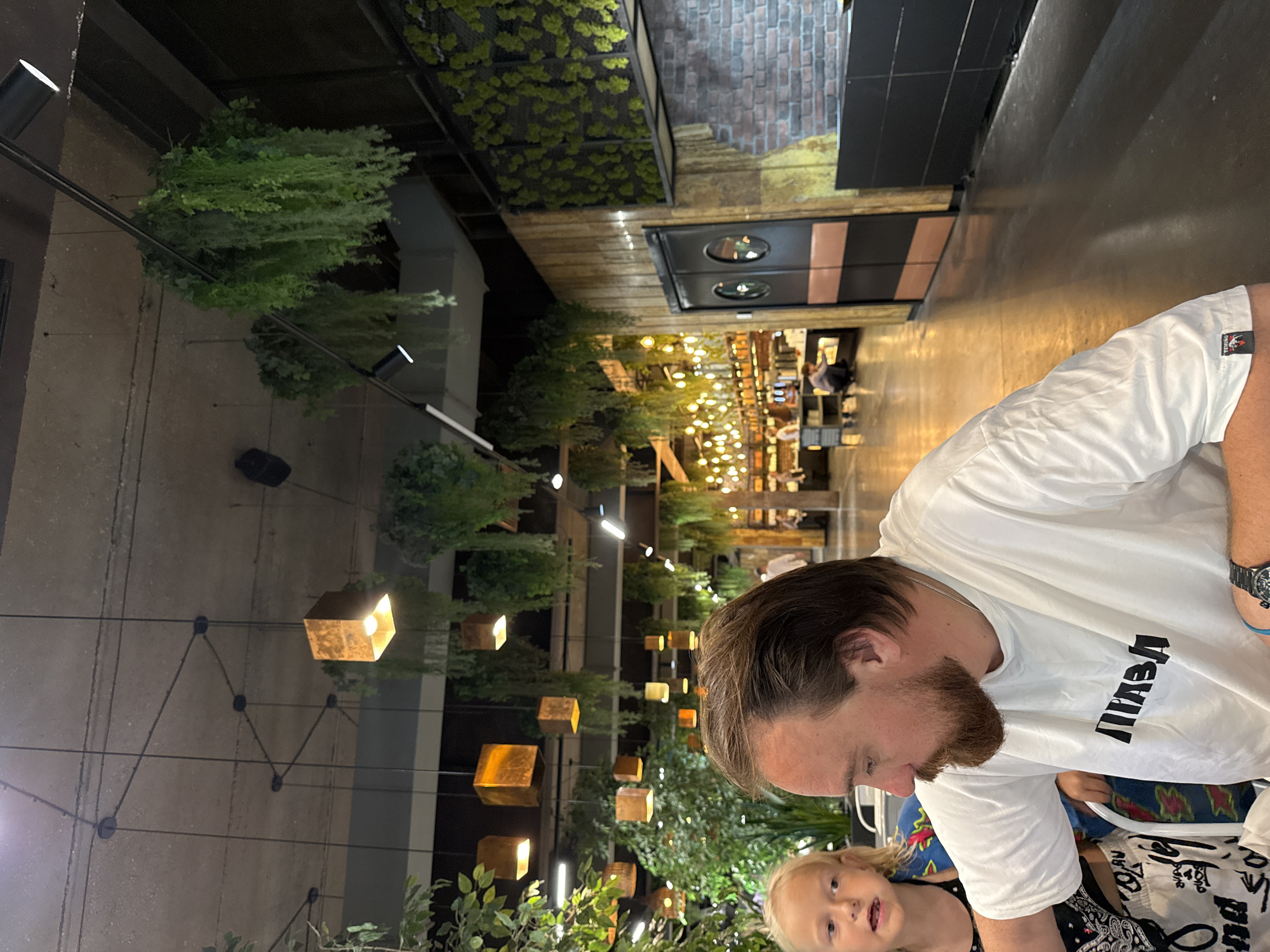 In the city of Yalta, Ukraine, a man with a beard and a white t-shirt with black Cyrillic lettering, and a young girl with blonde hair are seated at a table in a restaurant. The restaurant's interior features hanging plants, decorative lighting, and exposed concrete ceilings. In the background, other patrons are visible, as well as a counter area with shelves stocked with items. The overall atmosphere appears casual and contemporary.