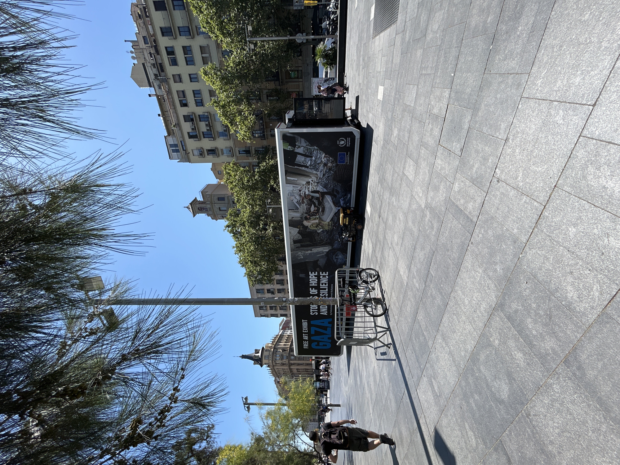 The image captures a street scene in Barcelona, Spain, featuring an outdoor art exhibit. In the foreground, a person wearing a black t-shirt and shorts with a backpack walks away from the camera on a paved plaza. To the right of the plaza is a large, dark-colored trailer serving as an art exhibit titled "FREE ART EXHIBIT GAZA STORIES OF HOPE AND RESILIENCE." The trailer's side displays images, and a small generator is positioned next to it. In front of the trailer, a metal barrier encloses two bicycles. In the background, several multi-story buildings line the street, and trees with green foliage are visible. The sky is clear and blue.