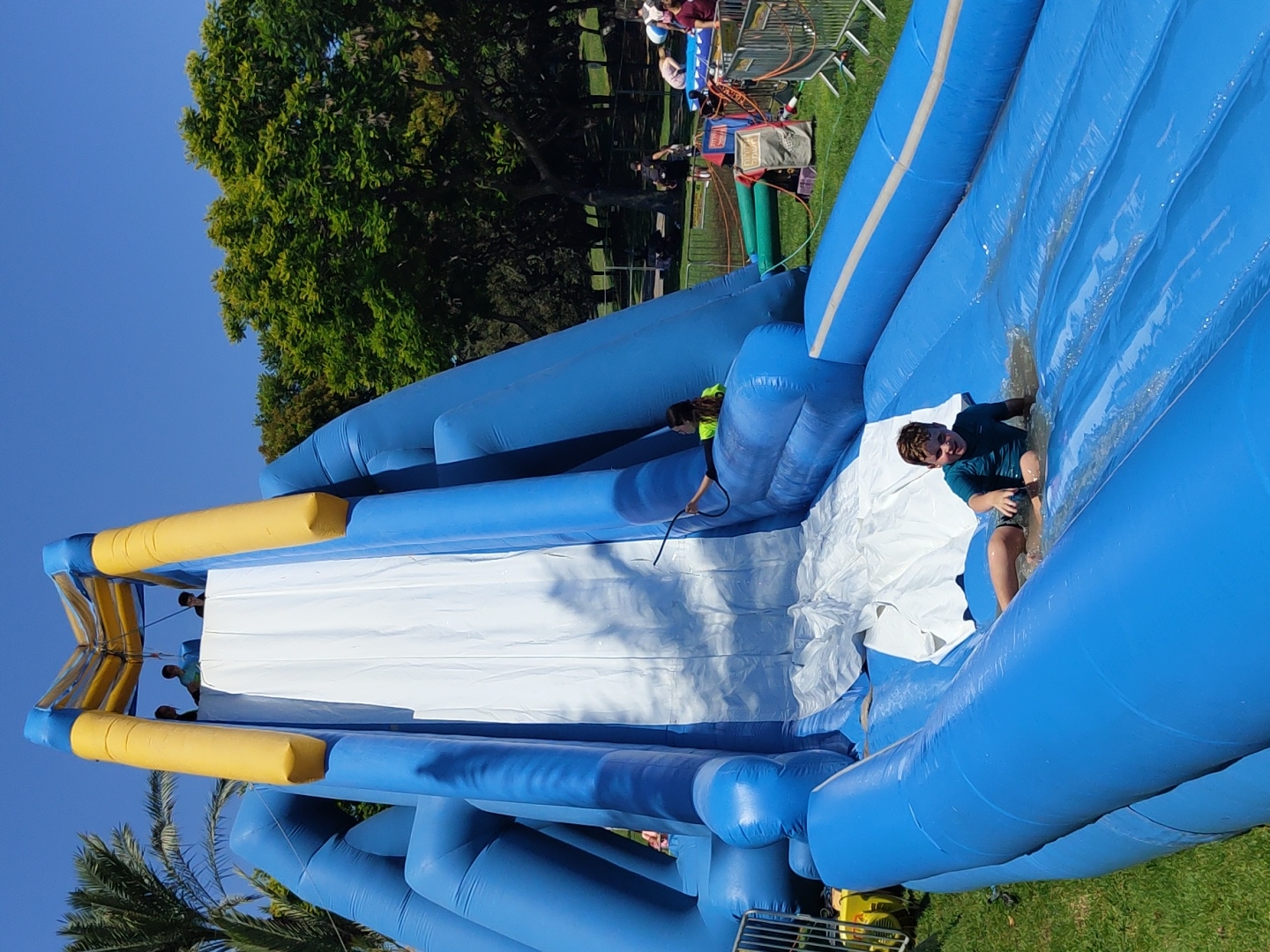 A young boy in a blue-green swimsuit is sliding down a large, inflatable blue water slide on a sunny day. He is at the bottom of the slide, sitting in a pool of water, with his legs extended forward. The water slide is multi-colored, with blue inflatable sections and a white sliding surface. In the background, other children and adults are visible, some near the top of the slide and others in the surrounding park area. Trees and a clear blue sky complete the scene. The event is likely taking place in Raanana, Israel, given the presence of palm trees and the general park environment.