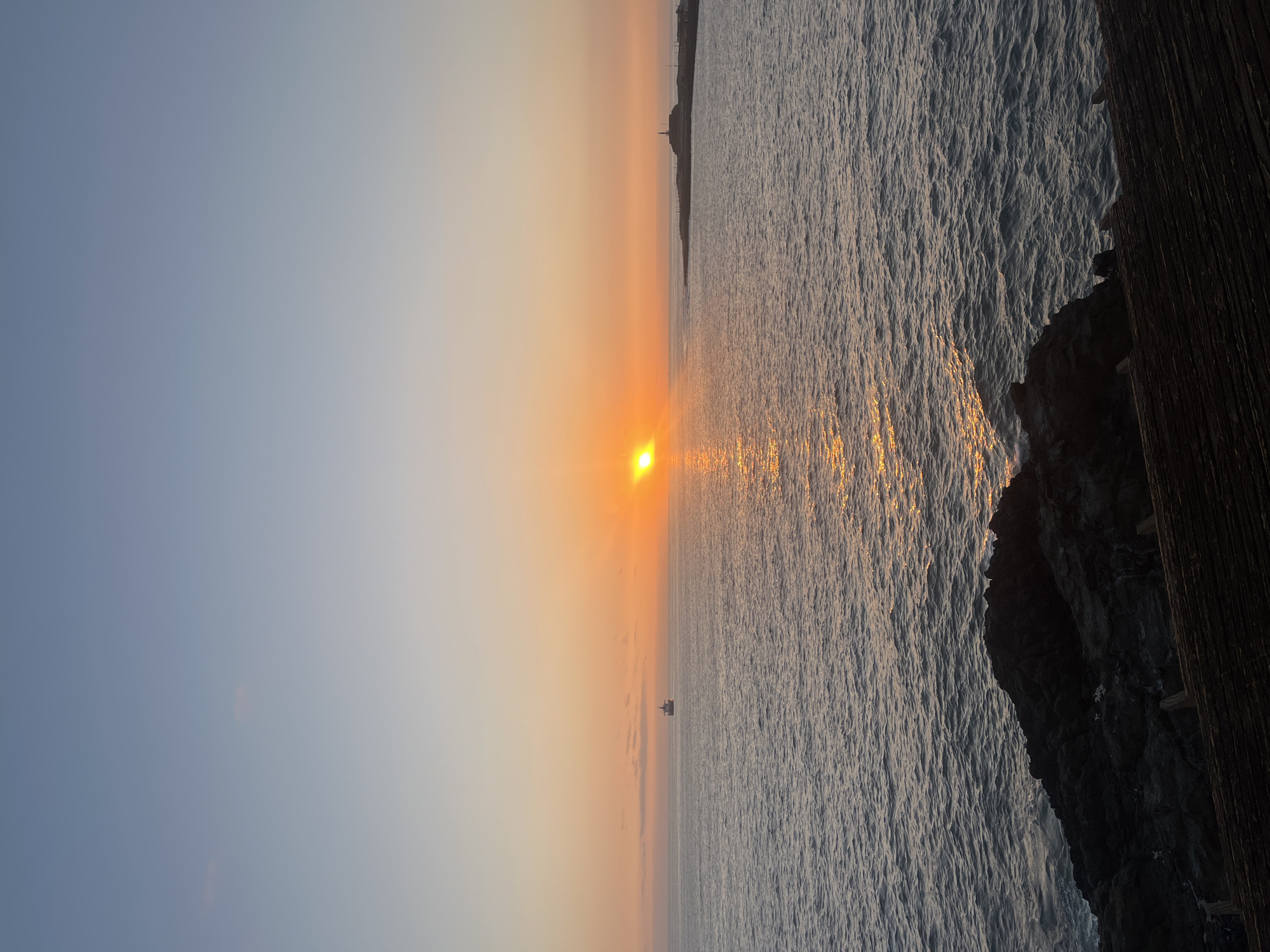 The image captures a sunset view in Arica, Chile. The sun is positioned near the horizon, casting a golden glow across the water and sky. A small boat is visible on the water. To the right, rocks and wood are visible. In the far distance, a building is visible atop the coastline.