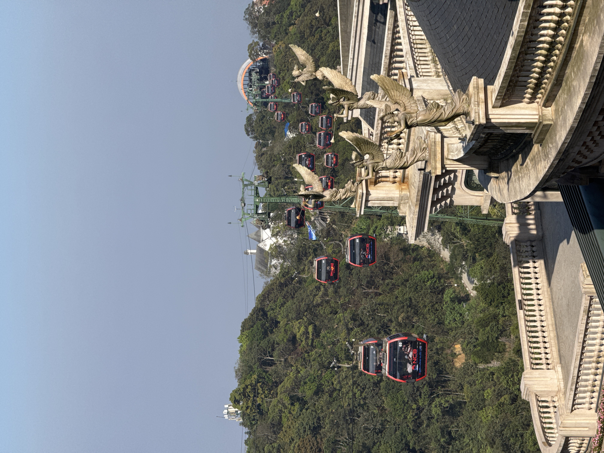 The image depicts a cable car system at Ba Na Hills, Đà Nẵng, Vietnam. In the foreground, a multi-tiered, stone-colored architectural structure with neoclassical balustrades is visible. Multiple statues of winged figures, resembling angels, are positioned on pedestals along these balustrades, many appearing to hold instruments. Partially visible dark-tiled, curved roofs extend below the balustrades.
Extending from the mid-ground towards the background, numerous red and black cable car cabins traverse a densely forested mountainside on a network of cables supported by green metal pylons. At least two cabins in the lower left contain visible passengers. Several cabins feature "NCB" and "NGAN HANG QUOC TE" branding, with one also displaying a "Red Bull" logo. Higher up the mountain, several white, European-style buildings are integrated into the dense forestation. In the upper right, a large arched structure, consistent with a cable car station, is situated on a peak. A prominent white communication tower is visible on a separate distant peak in the upper left. The sky above is clear and light blue.