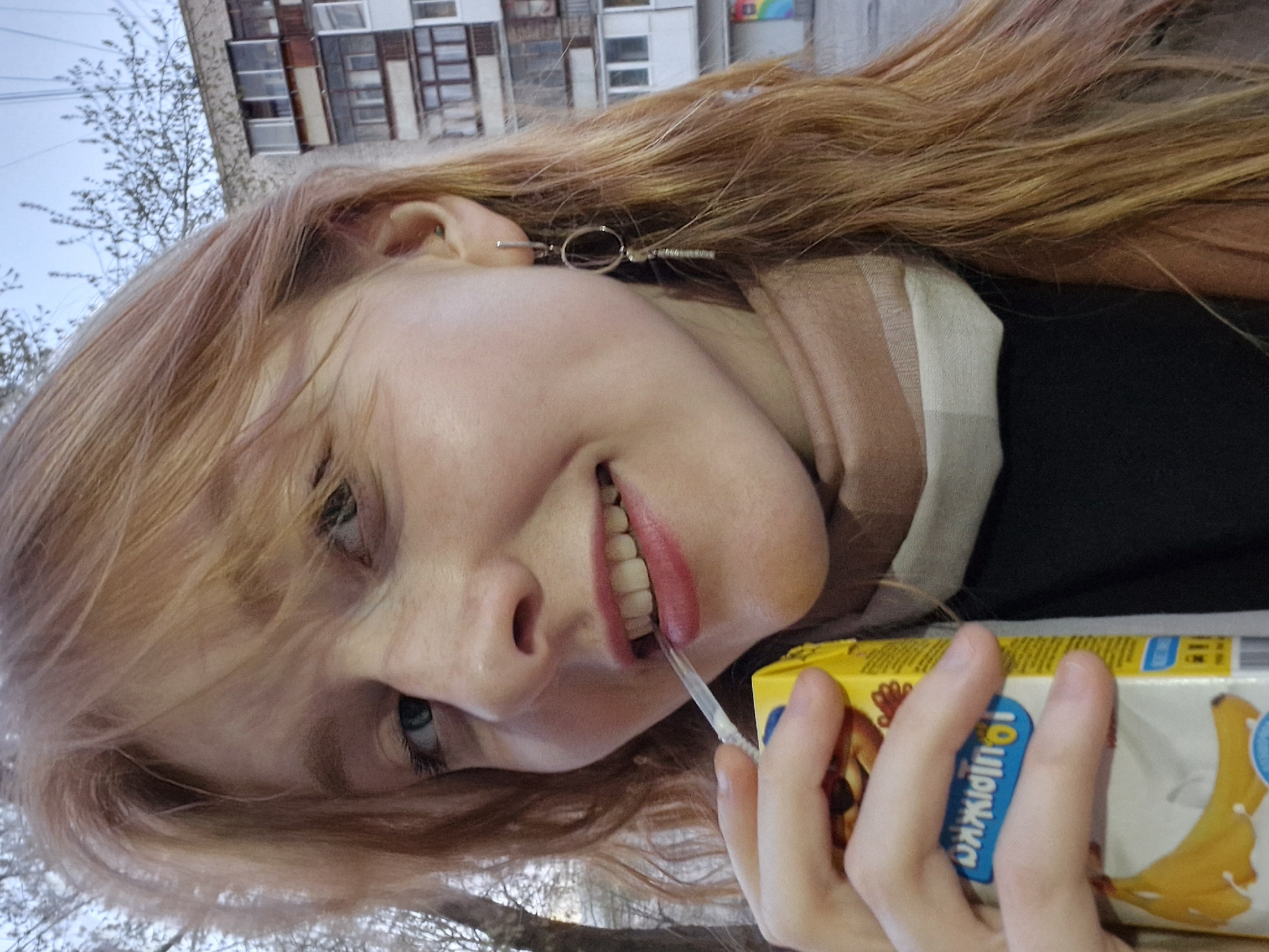 A close-up, angled outdoor portrait captures a young person, likely female, with long, reddish-blonde hair and light-colored eyes. She is smiling broadly, revealing her teeth, and appears to be enjoying a beverage from a juice box. Her lips are adorned with pink lipstick.
She is wearing a dark top and a light-colored scarf, possibly with a subtle plaid or striped pattern, around her neck. A distinctive earring, featuring a small hoop and a horizontal bar with tiny details, is visible on her right ear.
The juice box, held in her right hand, is primarily yellow and white. A prominent blue label on the side clearly displays the text "Садочек" (Sadochok), identifying it as a common juice brand in Russia. An illustration of a banana on the front confirms the flavor.
In the background, against an overcast sky, are the bare branches of trees, suggesting early spring or late autumn. Beyond the trees, a large, multi-story apartment building with numerous windows is visible, reflecting typical Soviet-era architecture found in cities like Yekaterinburg, Russia. The overall mood is cheerful and casual.