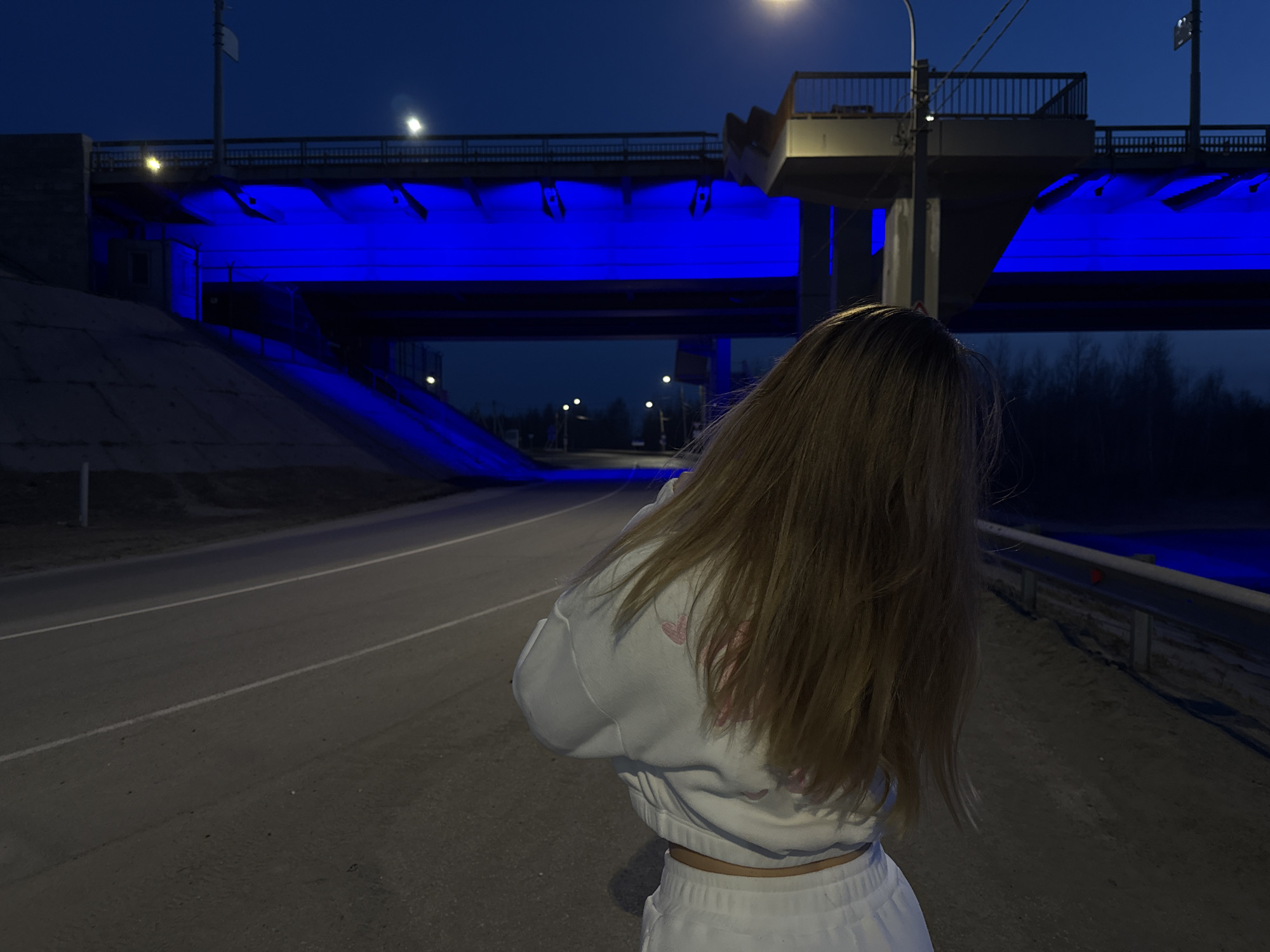 The image captures an outdoor night scene featuring a person and an illuminated bridge. In the foreground, a person is seen from behind, their long, light brown or blonde hair flowing over their back. They are wearing a white cropped sweatshirt with faint pink details on the back and matching white sweatpants or similar bottoms, revealing a sliver of their midriff. The person stands on the edge of an asphalt road, which stretches into the distance, marked by two parallel white lines.
The dominant feature of the background is a large bridge or overpass, strikingly illuminated from underneath with vibrant blue lights. These lights cast a blue glow on the underside of the bridge structure and partially on the ground beneath it, including a sloped concrete embankment to the left. Above the blue lights, the bridge features a sturdy railing. Several streetlights are visible, some on the bridge itself and others along the road ahead, providing patches of warm light against the dark, clear night sky. To the far right, faint outlines of trees can be seen in the distance, suggesting a more rural or undeveloped area beyond the immediate infrastructure. The road features a metal crash barrier along its right side. No visible text is present in the image.