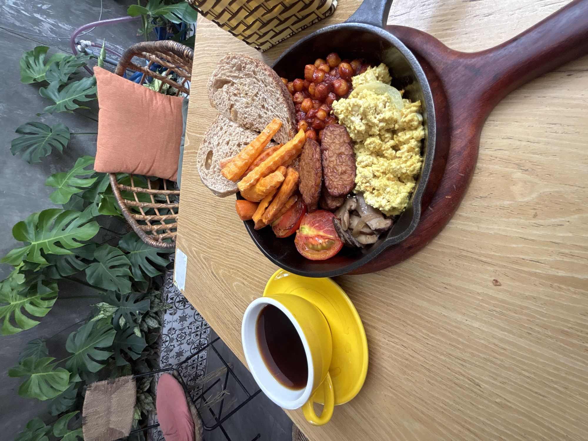 A close-up overhead shot shows a healthy breakfast on a light brown wooden table. In the foreground, a bright yellow cup and saucer hold dark coffee. To the right, a cast-iron skillet filled with a colorful vegan breakfast sits on a wooden board. The skillet contains scrambled tofu, baked beans, sliced tomatoes, sauteed mushrooms, tempeh strips, and sweet potato fries. Two slices of whole-wheat bread are placed on top of the food. In the background, lush green plants grow against a gray wall, with glimpses of rattan chairs and cushions. The scene appears to be in a cafe or restaurant in Da Nang, Vietnam.