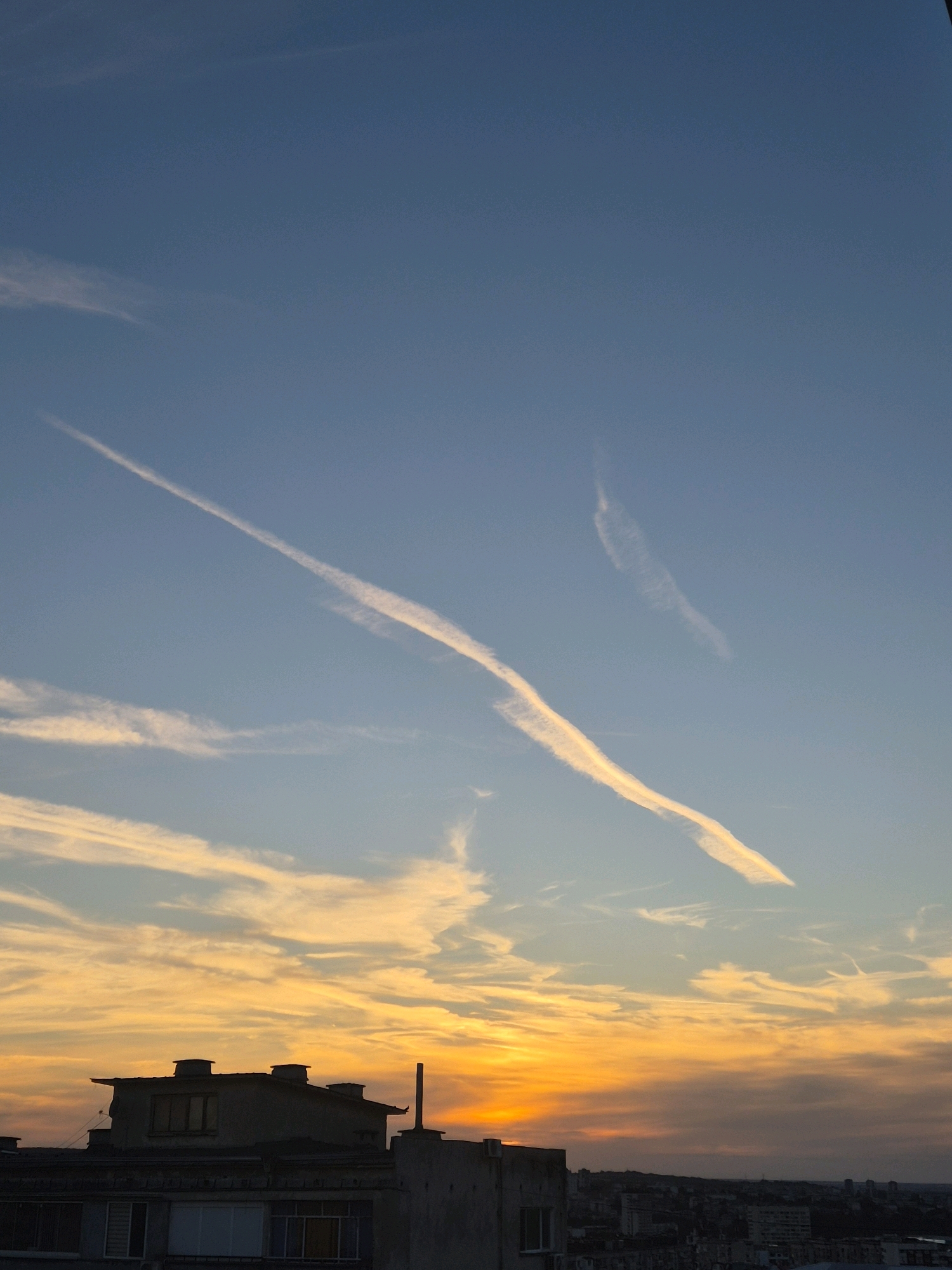 The image captures a sunset over a city. The sky is a gradient of blue at the top, transitioning to vibrant orange and yellow hues near the horizon. Two prominent contrails, likely from airplanes, stretch across the blue sky. One contrail is long and diagonal, while the other is shorter and more fragmented. Wispy clouds are scattered throughout, catching the warm light of the sunset. In the foreground, the silhouettes of buildings are visible against the colorful sky. These buildings appear to be of varied heights and architectural styles, suggesting an urban environment. The overall scene evokes a sense of twilight and the end of the day in a city.