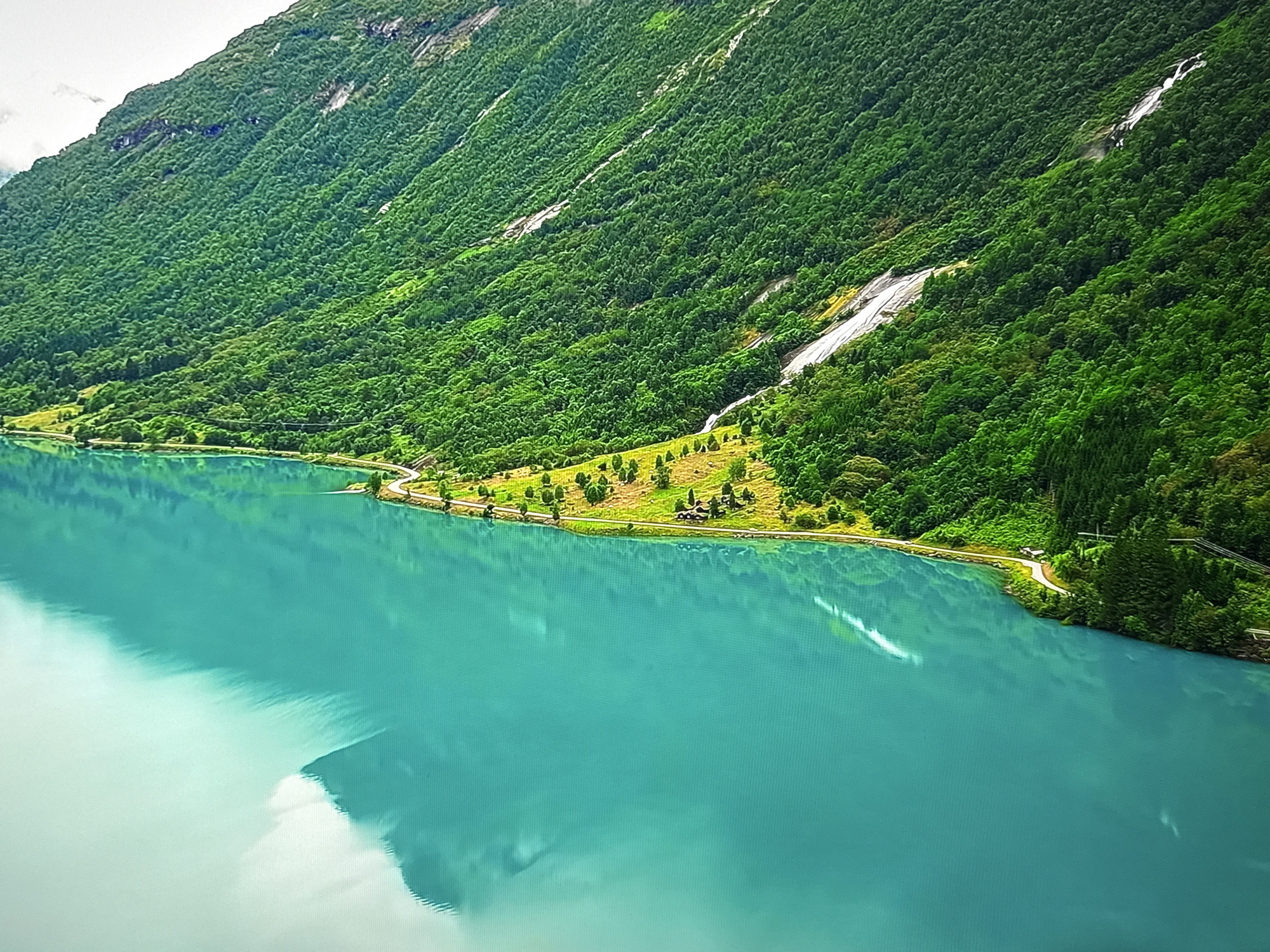 The image depicts a vast, tranquil body of turquoise water in the foreground, reflecting the sky and the surrounding landscape. The water stretches towards a verdant, tree-covered mountain that rises steeply from the shoreline. A thin, winding road is visible along the base of the mountain, near the water's edge, with scattered trees and small buildings nestled on a strip of land between the road and the mountain. The mountain itself is covered in dense green foliage, with some lighter streaks suggesting rock formations or waterfalls cascading down its slopes. The overall scene conveys a sense of natural beauty and serenity. This image is not located in the city of Burgas, Bulgaria.