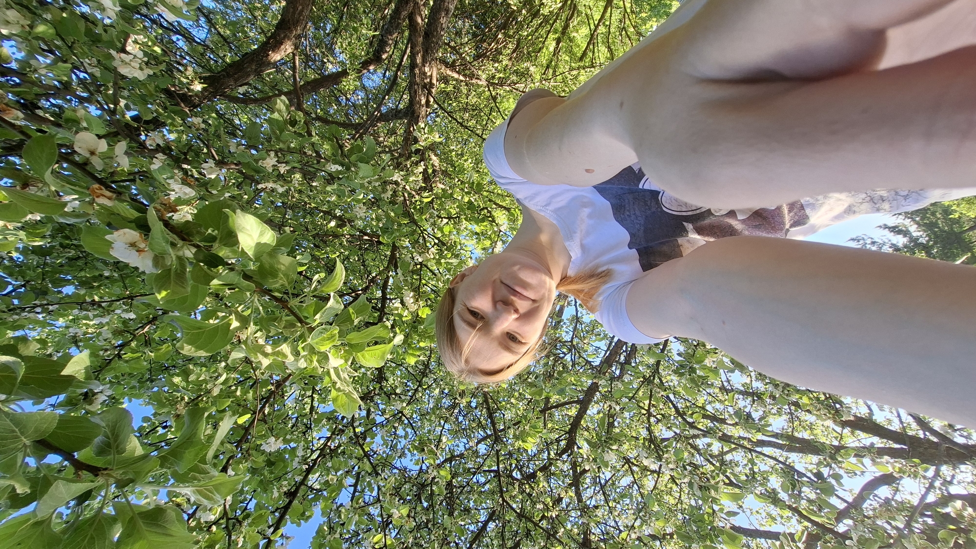 A selfie taken from a low angle shows a blonde woman in a white t-shirt standing beneath a tree with flowering white blossoms and green leaves. The blue sky is visible through gaps in the branches. The location is Большой Букор, Russia.