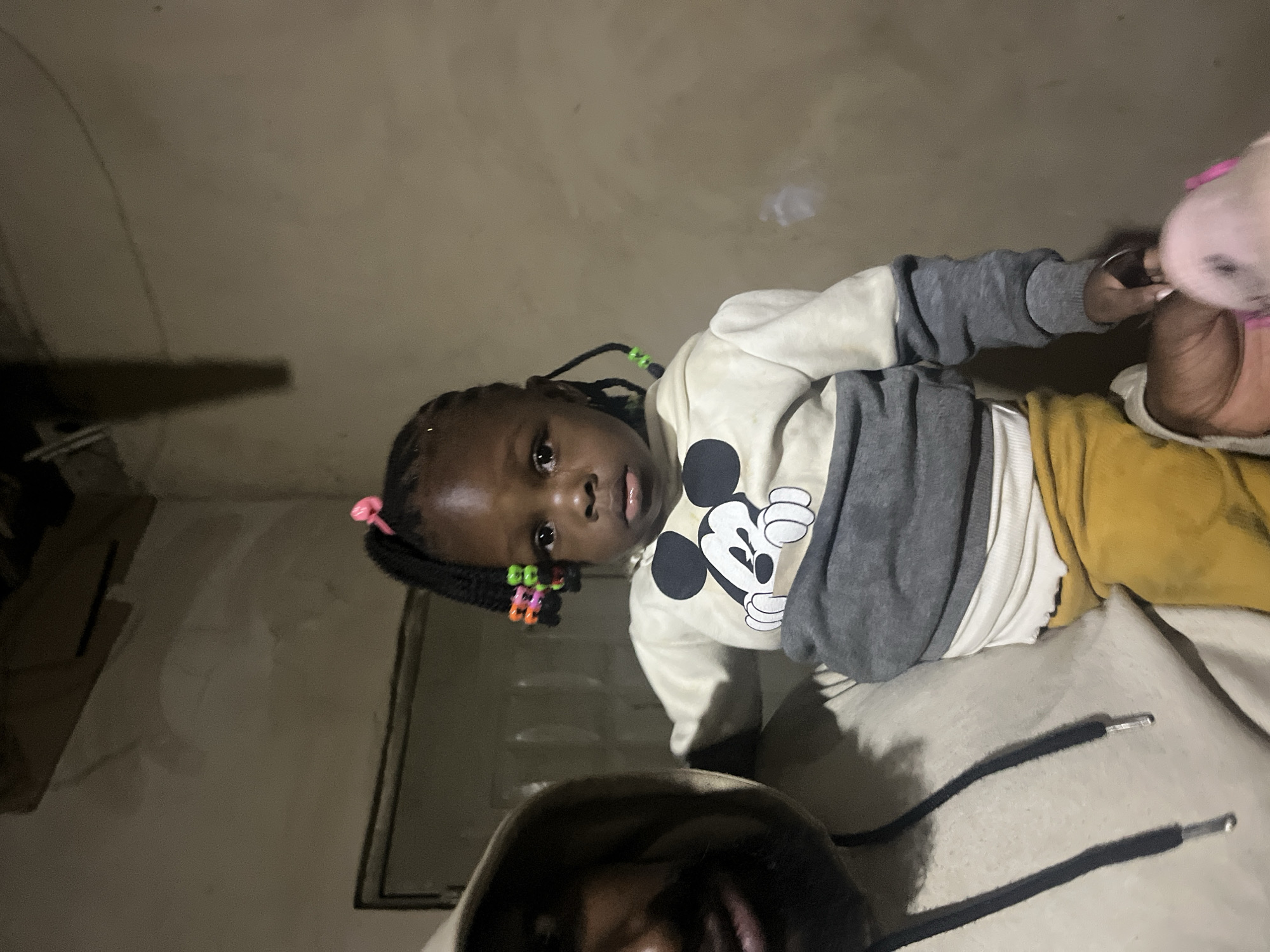 A person, partially visible on the left, holds a young Black child in their arms. The child has dark skin and braided hair adorned with colorful beads and a pink hair tie. The child is wearing a cream-colored hooded sweatshirt with a large Mickey Mouse graphic on the front and gray sleeves. The child is also wearing mustard-yellow pants. The background appears to be an indoor setting with beige walls and a dimly lit, unfinished ceiling.