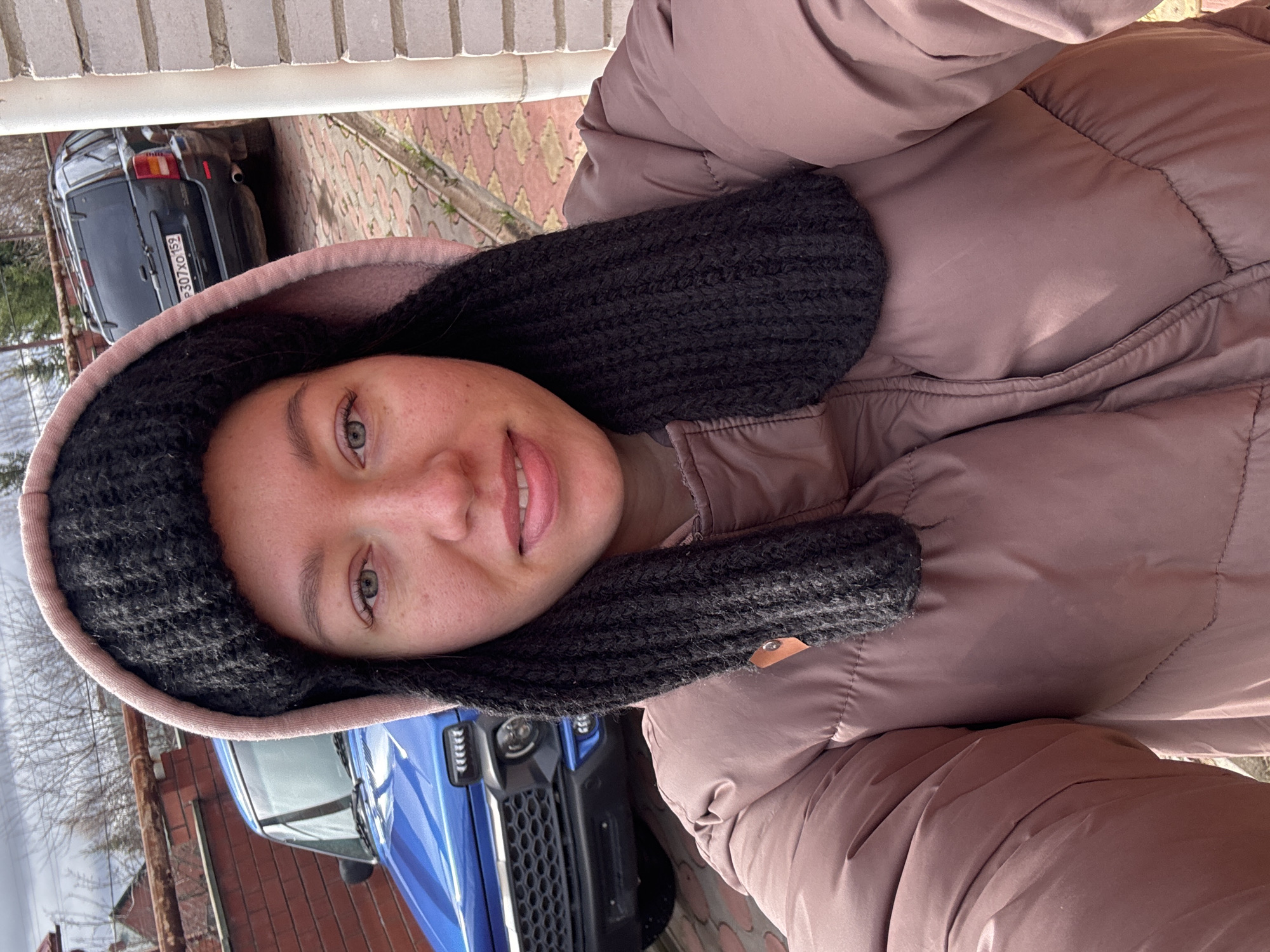 A woman wearing a puffy mauve jacket and a black knitted hood is taking a selfie. Part of a blue car is visible in the lower portion of the frame, with a brick building behind it. The rear of a dark-colored car with the license plate "P307XO 159" is seen in the upper left corner. The location appears to be a paved area, possibly a driveway or street. The setting might be in Ferma, Russia.