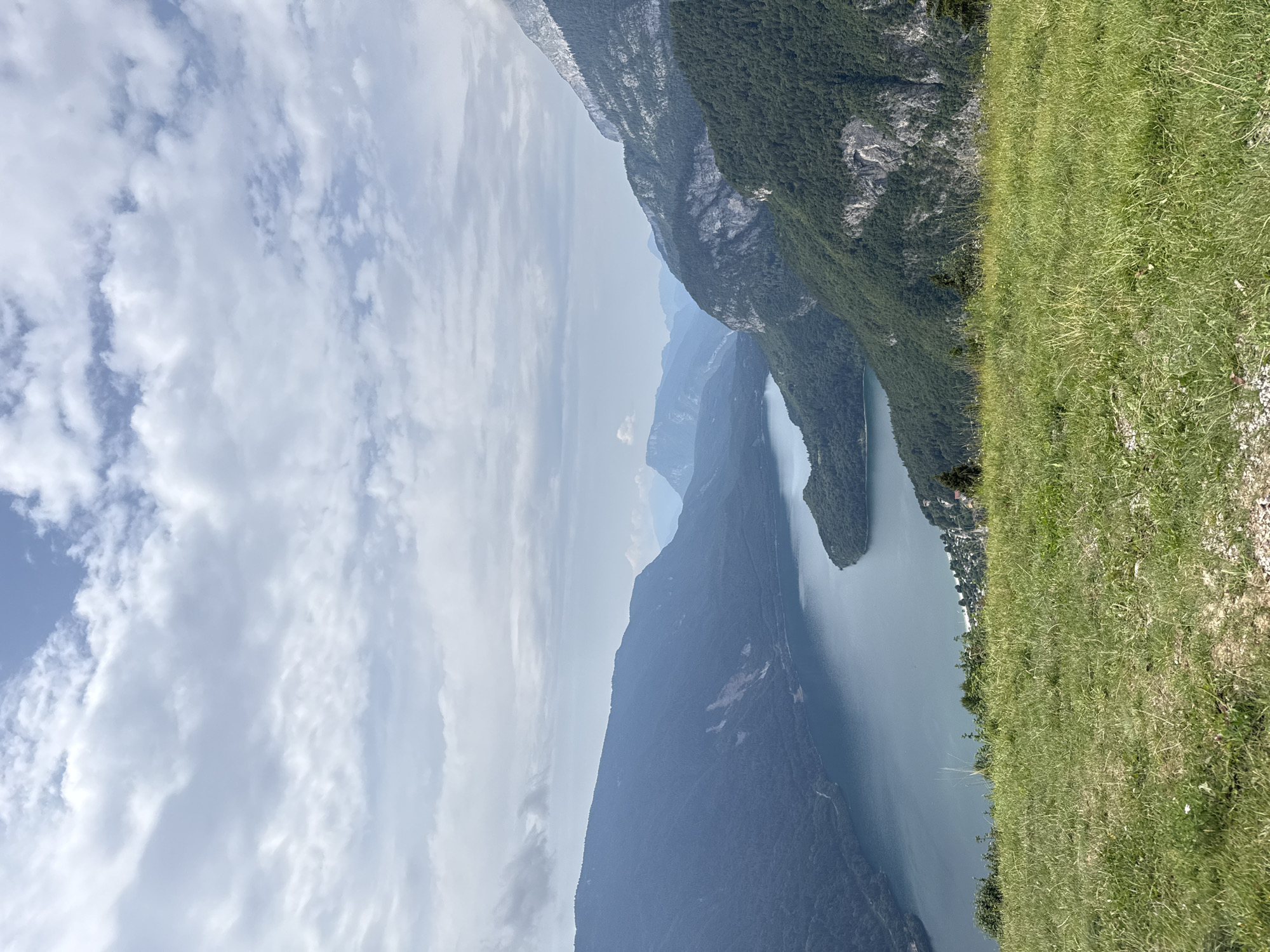 The image shows a panoramic view of Lake Molveno in Molveno, Italy. In the foreground, a grassy hill slopes down towards the lake. The lake itself is a deep blue, with calm waters reflecting the cloudy sky. Surrounding the lake are lush green mountains covered in trees. A small village or cluster of buildings is visible on the far shore of the lake. The sky is filled with scattered white clouds against a pale blue backdrop. The overall scene depicts a serene and picturesque natural landscape.