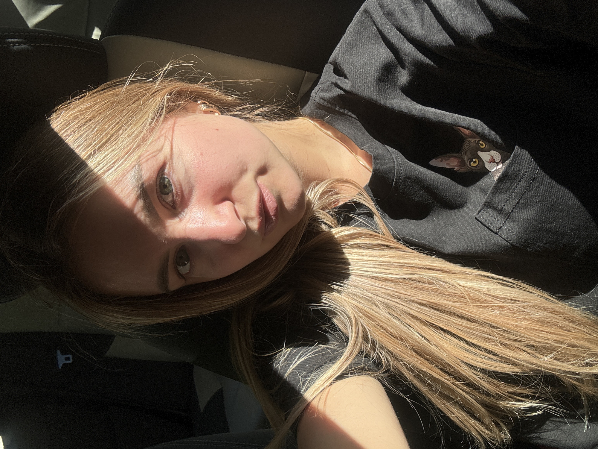 A young woman with blonde hair is sitting in the passenger seat of a vehicle, possibly a car. She is looking at the camera and her face is illuminated by sunlight. She is wearing a black t-shirt with a pocket that has a cartoon cat design on it. The background shows the interior of the car, including a headrest and a seatbelt. The lighting suggests it is daytime and the sun is shining through the window. There are no discernible landmarks or features that would place the image in the city of L'Essard, France.