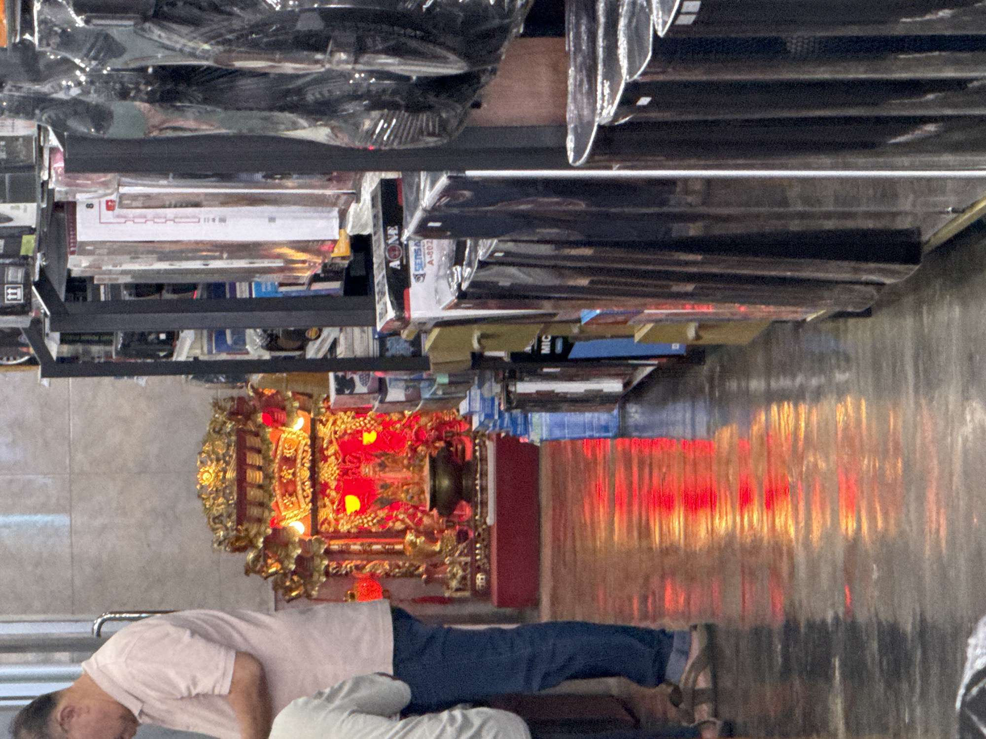 The image depicts an interior scene, possibly a shop, with a man in a light pink polo shirt and blue jeans standing on the left, facing away from the camera. Beside him, to the right, is a vibrant, ornate altar featuring a golden structure with red accents and illuminated by a bright red light. The altar has a dark urn or censer at its base. The floor is reflective, showing a strong red and orange glow from the altar's lighting. In the background and to the right, shelves are filled with various products, some of which are wrapped in plastic. The overall ambiance suggests a place of worship or a shop that incorporates religious elements. The location is identified as Ranong, Thailand.