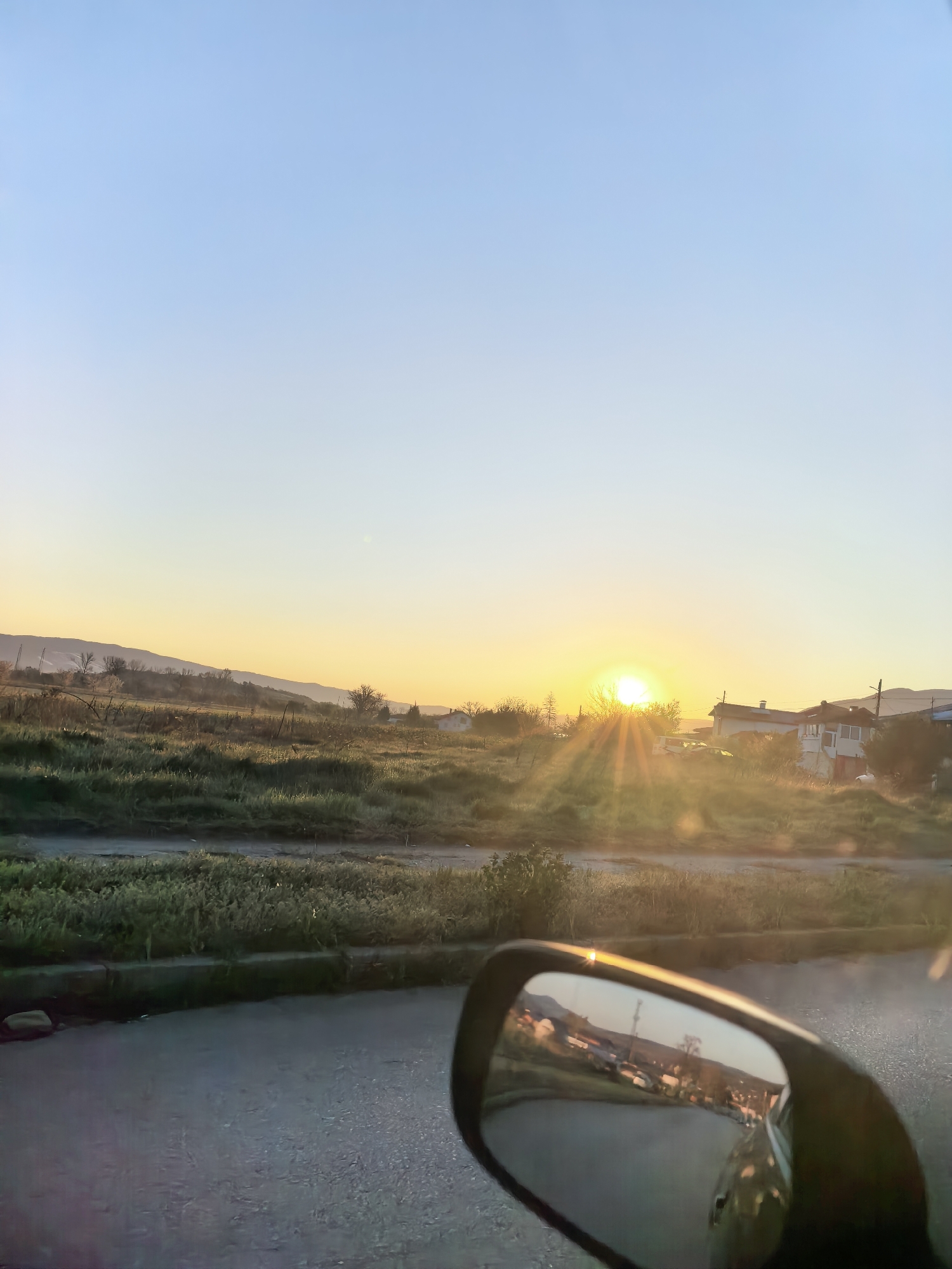 The image shows a rural landscape at sunrise or sunset, seen from inside a vehicle. A side mirror occupies the bottom right corner, reflecting a road and other vehicles. Beyond the roadside vegetation, there's a field, some houses, and hills in the distance, illuminated by the setting or rising sun.