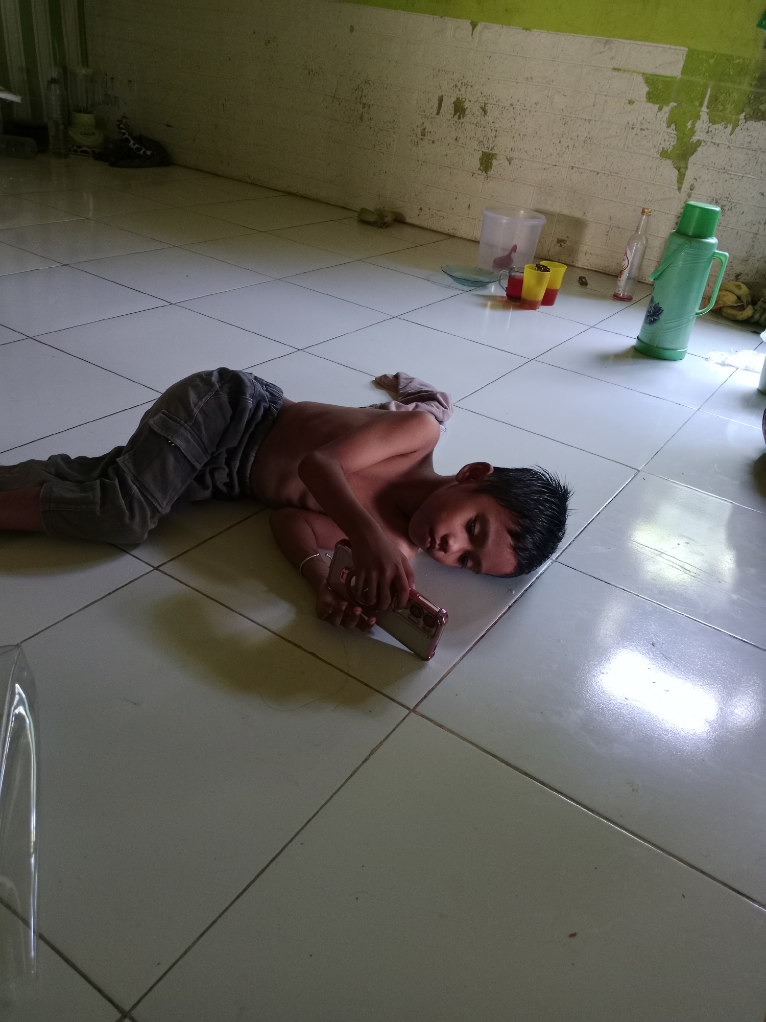 The image shows a young boy lying on a tiled floor. He is shirtless and wearing grey pants with cargo pockets. The boy is holding a smartphone in his right hand, and his eyes are closed, suggesting he is either asleep or deeply engrossed in the device. The floor is made of large, light-colored tiles. In the background, there is a wall with peeling paint, some plastic containers, cups, and a green thermos. The overall environment appears to be a humble dwelling. The scene is set in Lakea Satu, Indonesia.