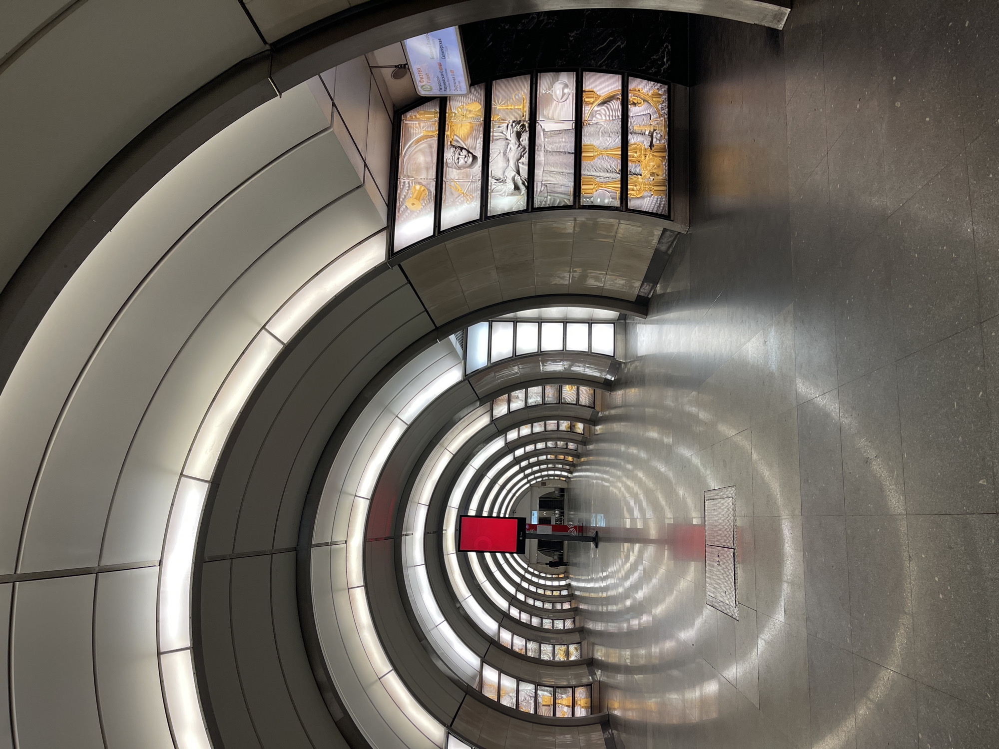 The image shows an underground station, likely a metro station, in a city identified as Unknown, Unknown. The architecture features a series of concentric arches forming a tunnel. On the left, the arches are solid and lit from within. On the right, the same arch structure appears to be reflected in the polished floor, creating a mirrored effect. On the upper-left part, the walls are covered with art, composed of gold and silver elements, inside of rectangular cases. Near the center of the image, a red electronic display screen is visible. The floor is made of large square tiles with a grey, speckled pattern. The tunnel recedes into the distance, converging into a vanishing point. There is no presence of any person inside of the metro station.