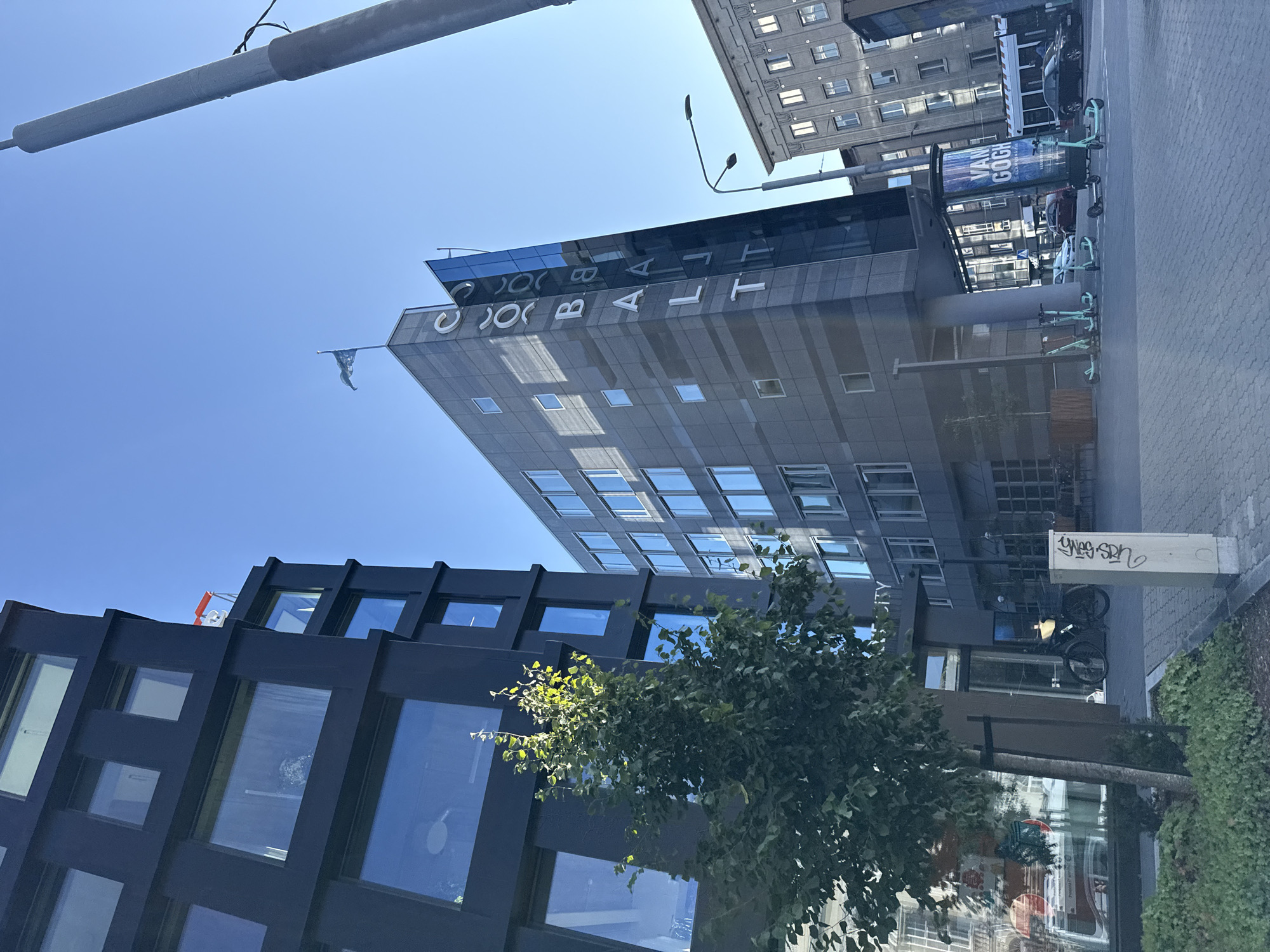 The image shows a street scene in Tallinn, Estonia, with a focus on modern architecture and urban elements. On the left, a dark, multi-story building with large windows dominates the foreground. To its right, a gray, angular building rises, featuring the word "COBALT" vertically inscribed on its side. A flag flies from the top of this building. In the background, older, lighter-colored buildings with multiple windows are visible. The street is paved with interlocking bricks, and several electric scooters are parked or being ridden. A bicycle is also present. A tall lamppost stands on the right side of the image. A poster with "VAN GOGH" and an illustration is displayed on a cylindrical structure. The sky is clear and blue. A small tree with green foliage is planted in the foreground. Graffiti is present on a utility box.