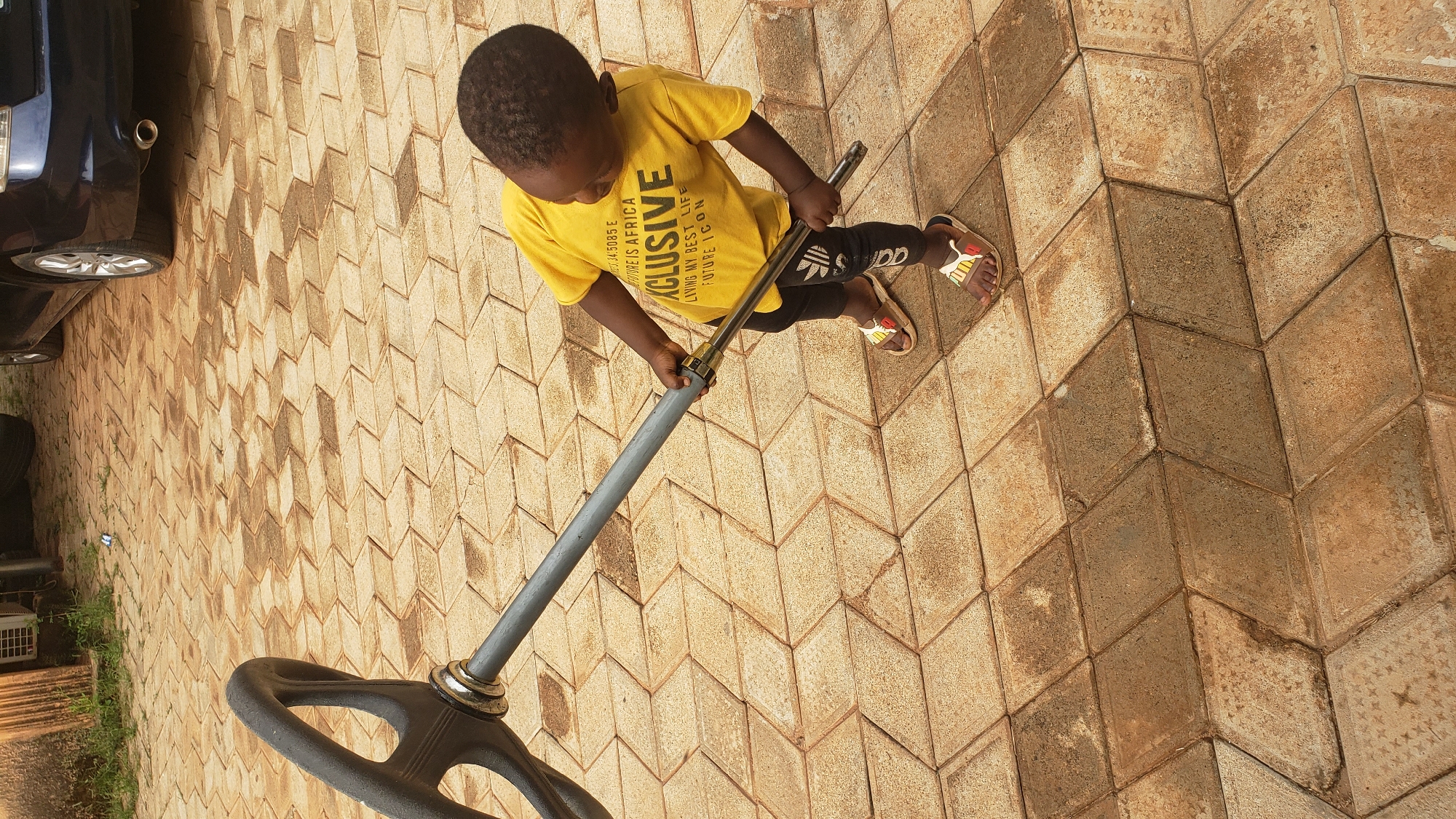 A young child, possibly a boy, wearing a yellow t-shirt with text that reads "AFRICA EXCLUSIVE" and black leggings with Adidas stripes, stands on a patterned, light brown paved area. He is holding onto a grey metal rod connected to a black steering wheel, appearing to be playing with a toy car or similar structure. The child is wearing sandals with a red and white design. The background shows a dark-colored car parked to the right and some overgrown green vegetation and a piece of equipment to the left. The scene is likely outdoors, possibly in a residential area of Sabon Tasha, Nigeria, given the paving pattern and the type of vegetation. The lighting suggests it is daytime, with shadows cast on the ground.