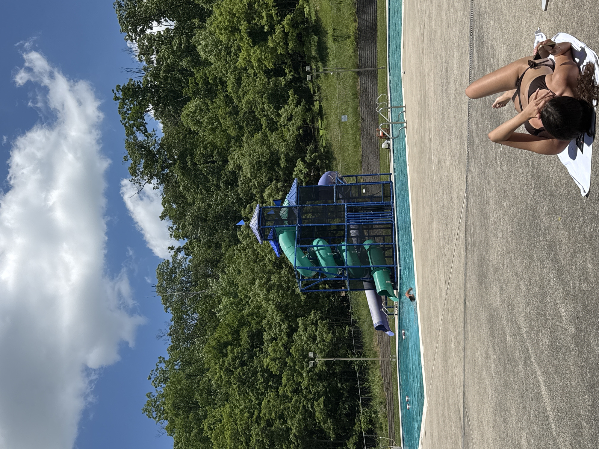 In Washington Township, a person is lying on a towel on a concrete surface near a swimming pool. The person is wearing a bikini and looking at their phone. The swimming pool is blue and has a water slide complex in the background. The water slide is tall and colorful, with green and purple slides. Behind the pool and slide, there is a dense forest with many trees. The sky is blue with scattered white clouds.