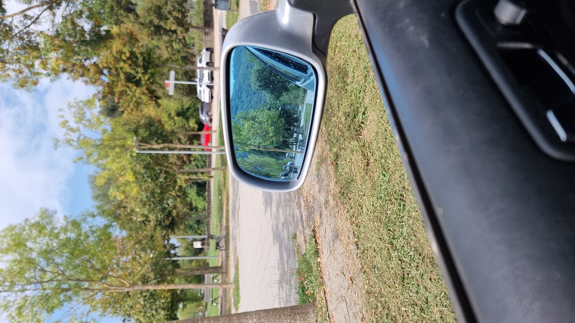 A side-view mirror of a car reflects a scene of trees, a paved area, benches, and cars. Outside the mirror, the foreground shows the car's interior door panel and grass. The scene appears to be in a park or recreational area with a backdrop of a wooded hillside. Based on the available data, this image was captured on July 23, 2023, at approximately 12:41 PM in Sant Joan les Fonts, Spain.