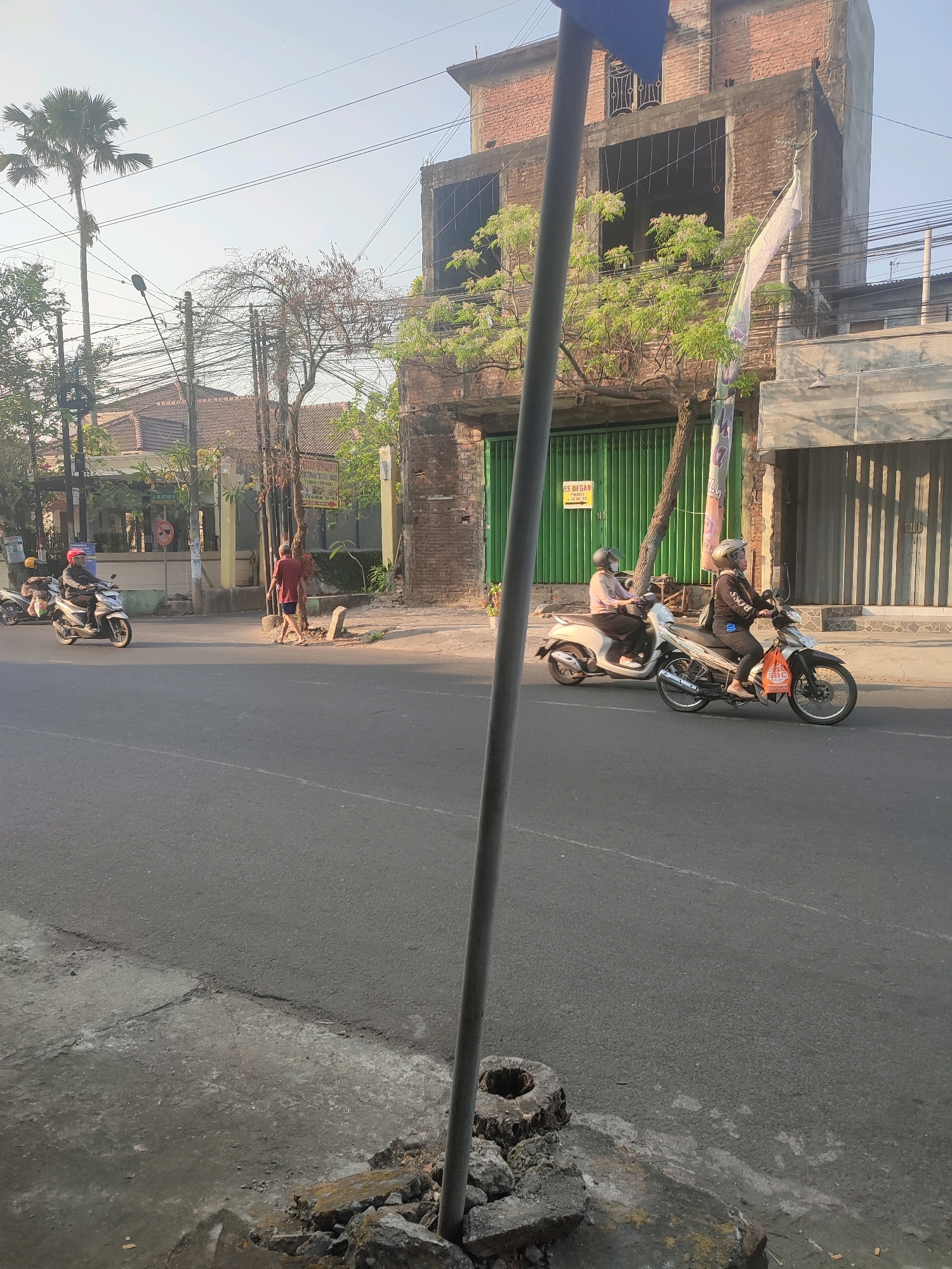 The image depicts a street scene in Nganjuk, Indonesia, under a clear, bright sky, suggesting it is either early morning or late afternoon due to the long shadows. In the foreground, a gray pole interrupts the view of the street, which is paved with asphalt and has a cracked concrete sidewalk on the left. A small pile of rubble and a tree stump are visible near the base of the pole.
Further down the street, two individuals are riding on separate motorcycles. The motorcycle on the left has a driver wearing a red helmet, and a passenger behind them with a yellow helmet. The motorcycle on the right has a driver wearing a helmet, and the motorcycle is carrying a patterned bag on its front. Both motorcycles are moving in the same direction.
Across the street, a building with exposed brickwork and unfinished upper floors stands. A green metal gate is visible in front of this building. To the left of this building, a smaller residential building with a tiled roof can be seen. A person in a red shirt and dark shorts is walking away from the camera on the sidewalk in front of the residential building. Several trees, including a tall palm tree, are present along the street. Electrical wires are strung overhead, supported by poles. A white flag is attached to the side of the brick building, fluttering in the breeze. A sign with text in Indonesian is also visible on the green gate.