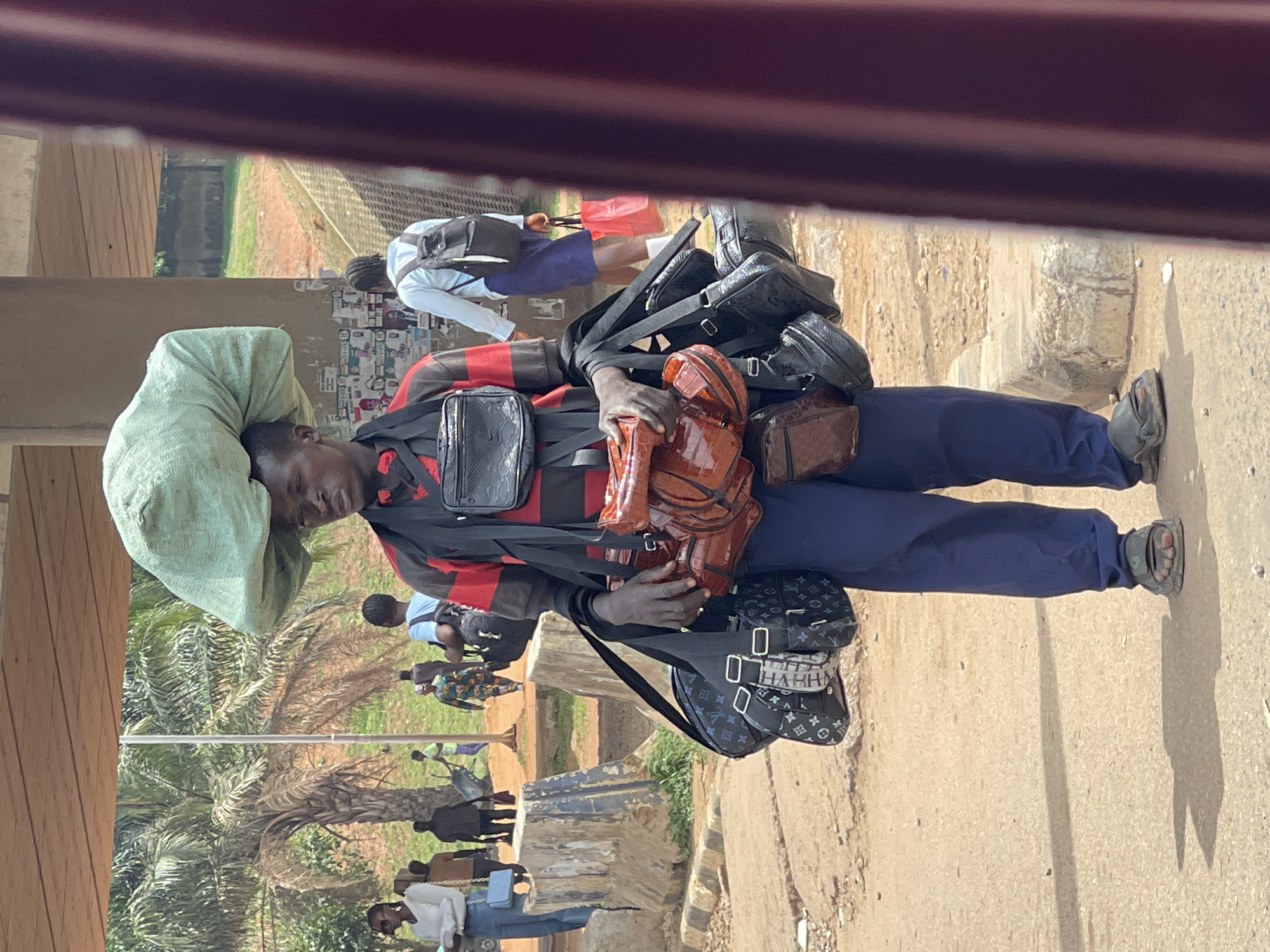 A man is standing on a sidewalk in what appears to be Abuja, Nigeria, with a large green bag on his head and several purses and bags hanging from his body. He is wearing a long-sleeved shirt with horizontal red and black stripes and navy blue pants. He is also wearing sandals. The sidewalk is made of concrete, and the ground appears to be sandy. In the background, there are other people and trees. There is a building in the foreground partially obstructing the view.