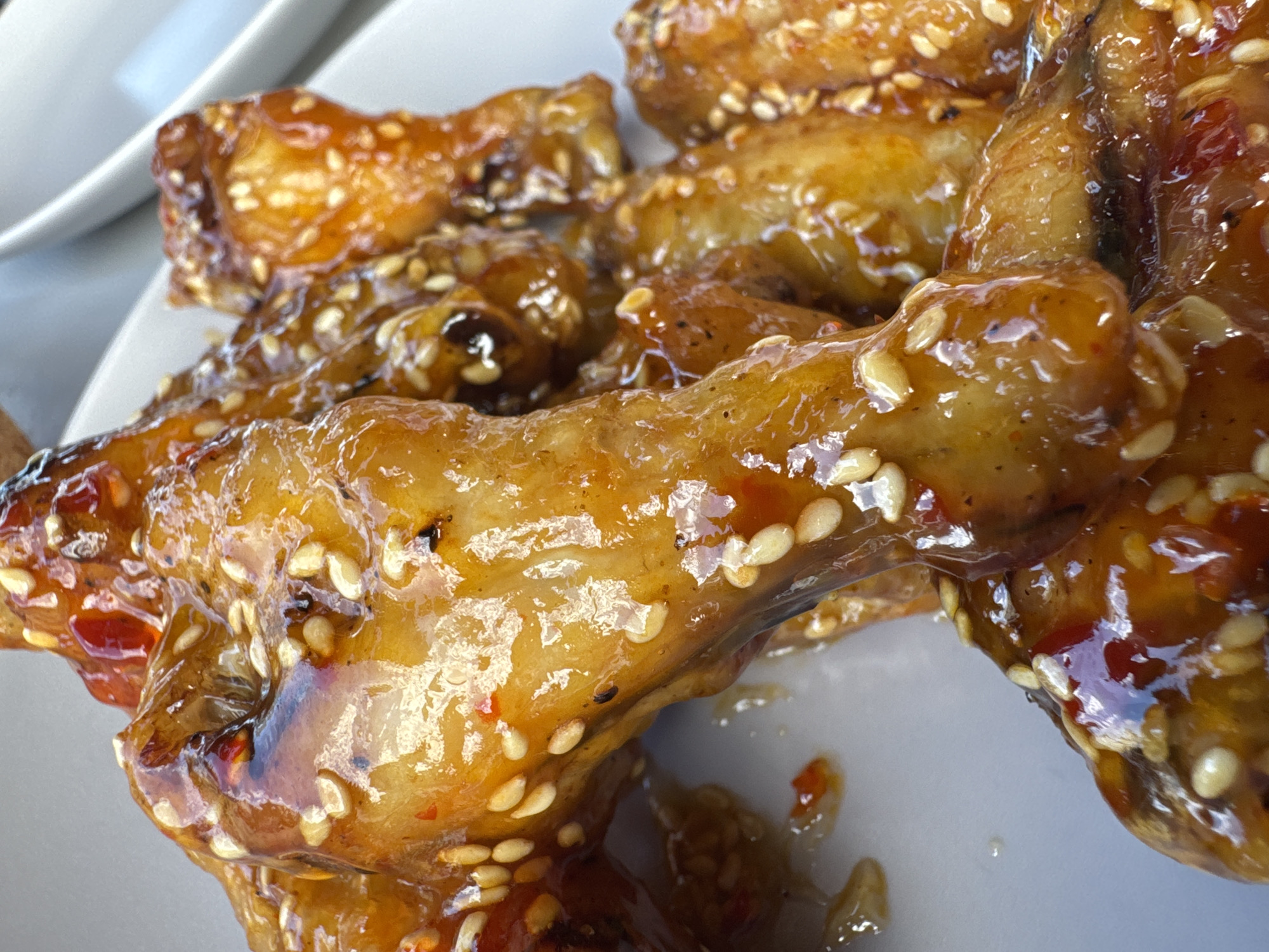 The image shows a close-up of cooked chicken wings covered in a glossy, dark orange sauce, garnished with white sesame seeds. The wings are piled on a white plate. Traces of sauce are visible on the plate. The location is not discernible from the image.
