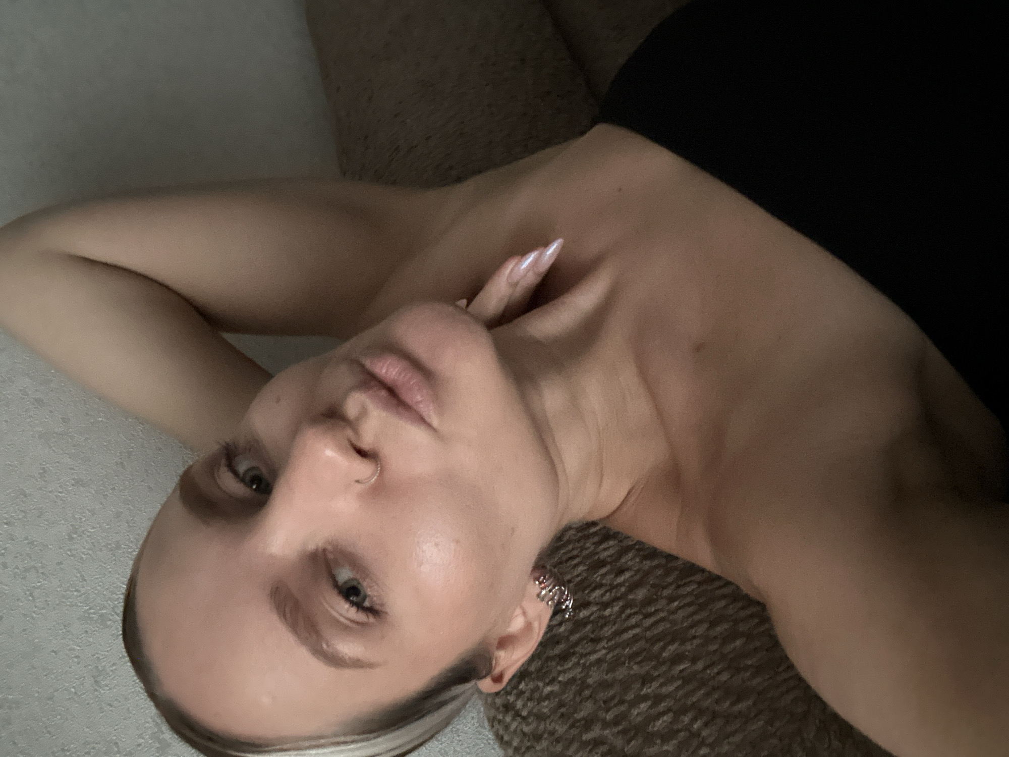 The image shows a close-up of a young woman lying down, with her head resting on her arm and her hand touching her neck. She is wearing a black strapless top and has a nose ring and hoop earrings. Her eyes are open, and her lips are slightly parted. The background appears to be a textured wall and a couch. The lighting is dim, creating a soft, intimate atmosphere. There is no identifiable information that places this image in Esenyurt, Turkey.