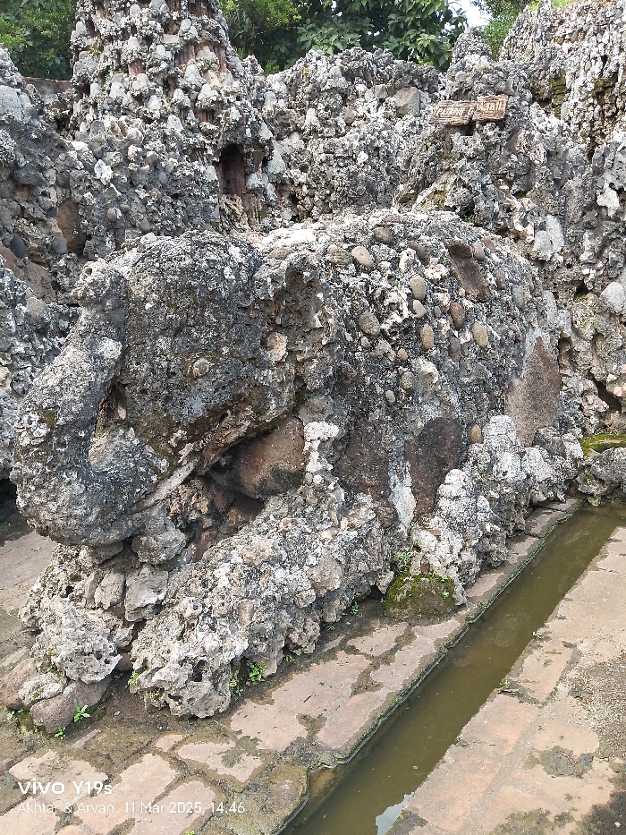 The image shows a large, grey, rocky structure, possibly a man-made cave or decorative rock formation, at the Keraton Kasepuhan palace in Cirebon, Indonesia, located at the given coordinates. The rock is formed from smaller stones and pebbles embedded in a matrix of cement or mortar. A long, narrow channel filled with murky water runs along the bottom of the image and alongside the base of the rocky structure. The channel appears to be made of several flat stones laid in a line. Vegetation grows sparsely around the edges of the water channel and at the base of the rock formation. There are no people present in the scene.