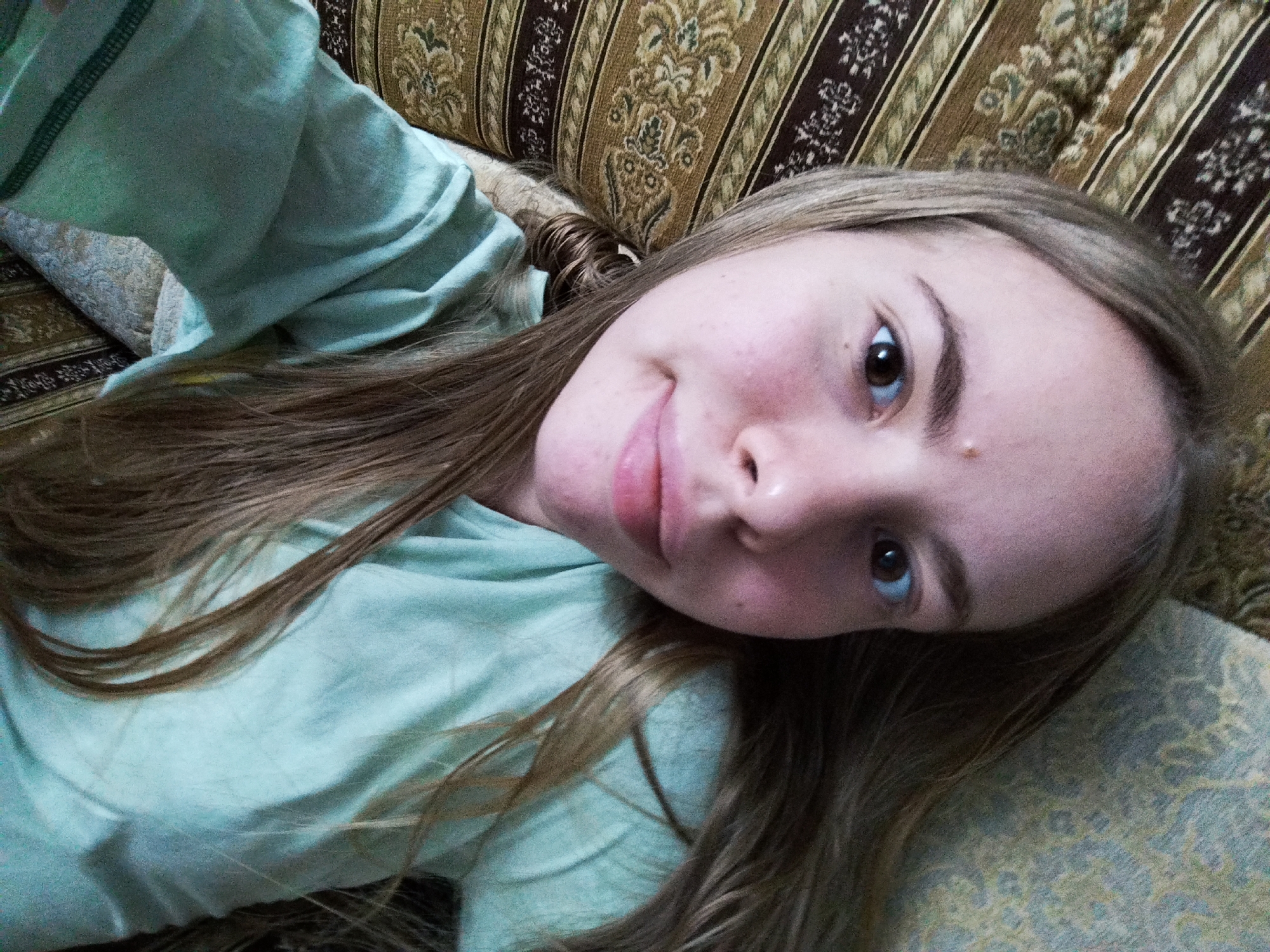 A young person with long brown hair and light skin is lying on a patterned couch. They are wearing a light blue t-shirt and looking up at the camera, with their head tilted to the side. The couch has a textured, striped pattern in shades of brown, gold, and cream. The lighting is dim, creating shadows on the person's face and hair. The background is out of focus, emphasizing the subject.