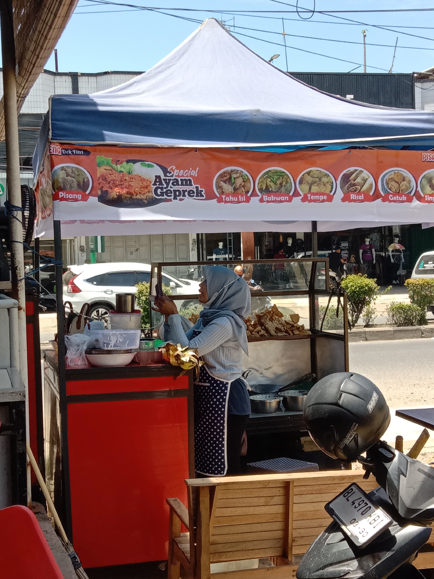 The image shows a street food vendor in Lhokseumawe, Indonesia, operating from a small, mobile stall under a white tent with a blue trim. The banner above the stall advertises "Special Ayam Geprek" along with other dishes, including "Pisang," "Tahu Isi," "Bakwan," "Tempe," "Risol," and "Getuk."
A woman wearing a light blue hijab and a polka-dot apron is standing at the stall, apparently looking at her mobile phone. She is positioned near a collection of bowls and cooking utensils. The stall itself is red with black trim, and a glass display case showcases various fried foods.
In the background, pedestrians wearing face masks walk on the sidewalk, and vehicles pass by on the road. A black motorcycle is parked in the foreground, bearing the license plate "BL 4970 KBI" with an expiration date of "03-30." The motorcycle has a black helmet hanging on it. A wooden bench is also visible, situated next to the stall.