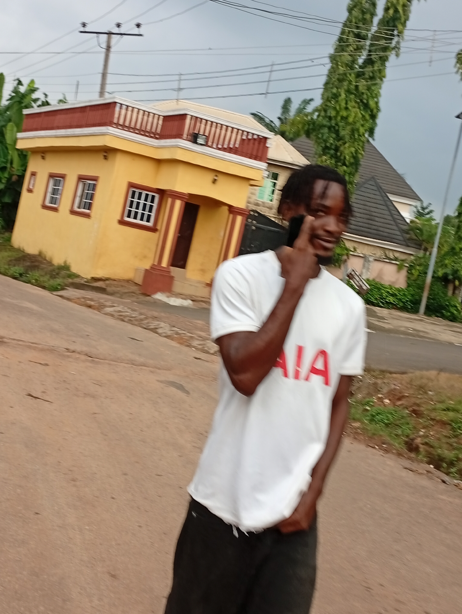 A young Black male stands in the foreground, facing slightly to his right and looking towards the camera. He is wearing a white t-shirt with red lettering that appears to spell out "AIA," and dark pants. His right hand is raised to his face, with his index finger touching his cheek, and he is holding a black mobile phone to his ear. He has dark, short hair.
Behind him, to the left, is a vibrant yellow, two-story building with a red-trimmed roof and white railings on the upper level. The building has multiple windows with white grids. A dark-colored door is visible on the ground floor. To the right of the yellow building, a large, dark-tiled roof of another building is visible. Tall, slender trees with dense green foliage rise behind the buildings. Overhead, power lines stretch across a cloudy sky. The ground in front of the buildings is a reddish-brown paved surface, suggesting a road or pathway. The overall scene depicts an outdoor environment in what appears to be a residential area, likely in the city of Nsugpe, Nigeria, given the architectural style and vegetation.