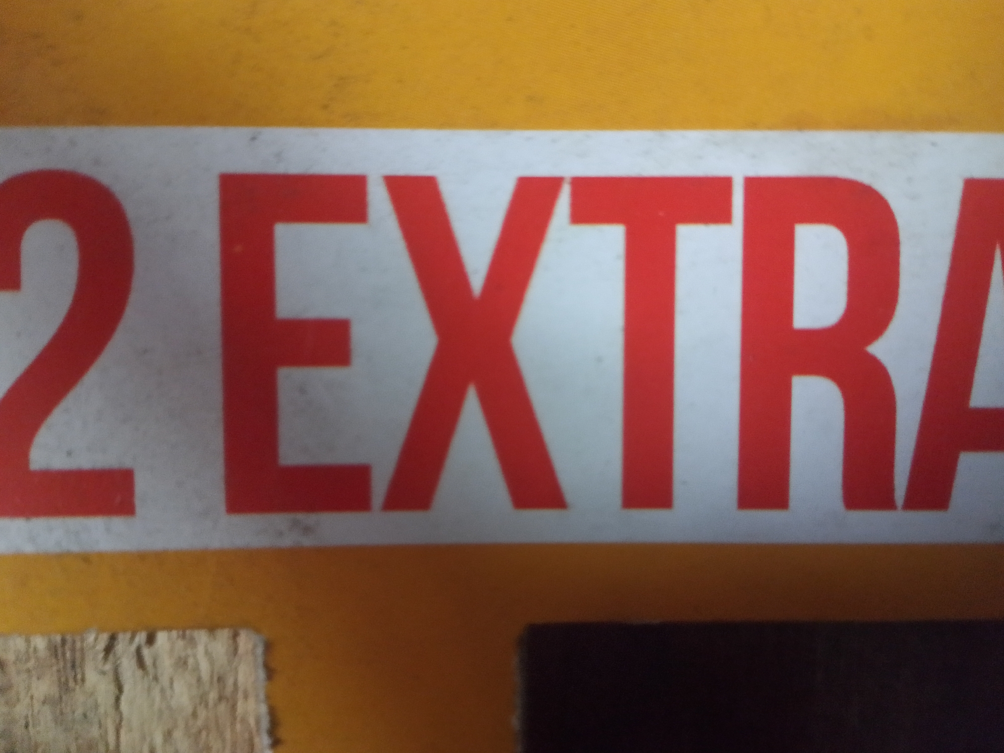The image shows a close-up of a white sticker with red lettering on a yellow background. The visible text reads "2 EXTRA". The sticker is partially obscured by what appears to be a piece of wood on the left and a dark, possibly black object on the bottom right. The overall scene suggests an outdoor or industrial setting given the textured yellow background and the presence of the wood. There are no people or specific landmarks visible to indicate a precise location within Mukapayung, Indonesia, beyond the general suggestion of a sticker being applied to a surface.