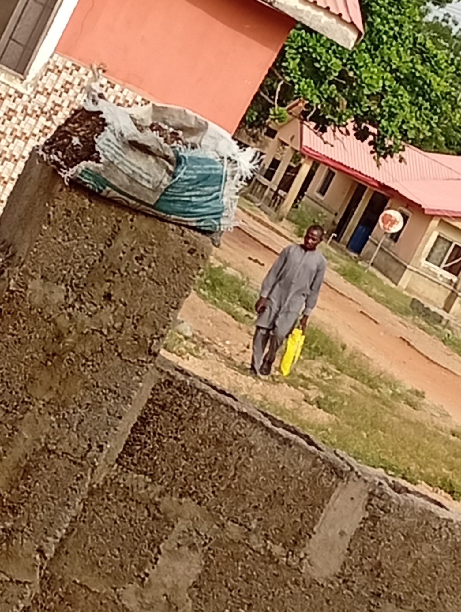The image captures an everyday scene in Adankolo, Nigeria, featuring a man walking along an unpaved path. In the foreground, on the left, a textured, brownish concrete or stone wall occupies a significant portion of the frame. On top of this wall rests a large, partially torn woven sack, in shades of white and blue/green, filled with dark, granular material, possibly soil or waste.
In the mid-ground, a man of medium build is seen walking towards the right side of the frame. He is dressed in a simple, long-sleeved grey traditional outfit, consisting of a tunic and trousers, paired with dark shoes. He carries a bright yellow plastic bag in his right hand. His expression is neutral, almost a slight smile, as he looks towards the camera.
The background reveals several buildings with distinct red corrugated roofs, characteristic of local architecture. One building in particular, located behind the man, has a prominent blue door and a white satellite dish with reddish markings mounted on a pole nearby. The ground is an uneven reddish-brown dirt path, interspersed with patches of green grass and some vegetation. Lush green trees and bushes are visible behind the structures, suggesting a green environment. The scene is bathed in natural daylight, indicating either mid-morning or late afternoon. No discernible text is visible on any objects or signs within the image. The overall impression is of a quiet, routine day in a local community.