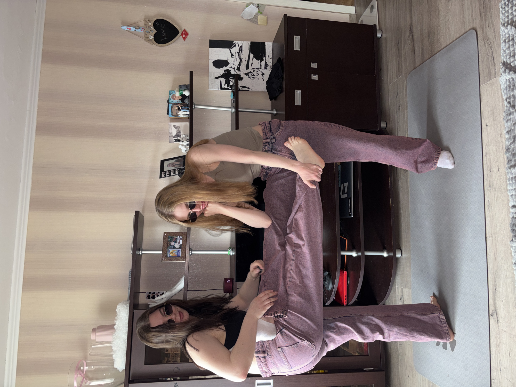 The image shows two women performing a yoga-like pose in what appears to be a residential room. Both women are wearing sunglasses and pink/purple trousers. One woman has blonde hair, and the other has dark hair. The setting includes a television stand, shelves with various objects, and a yoga mat. The location is identified as Perm, Russia.