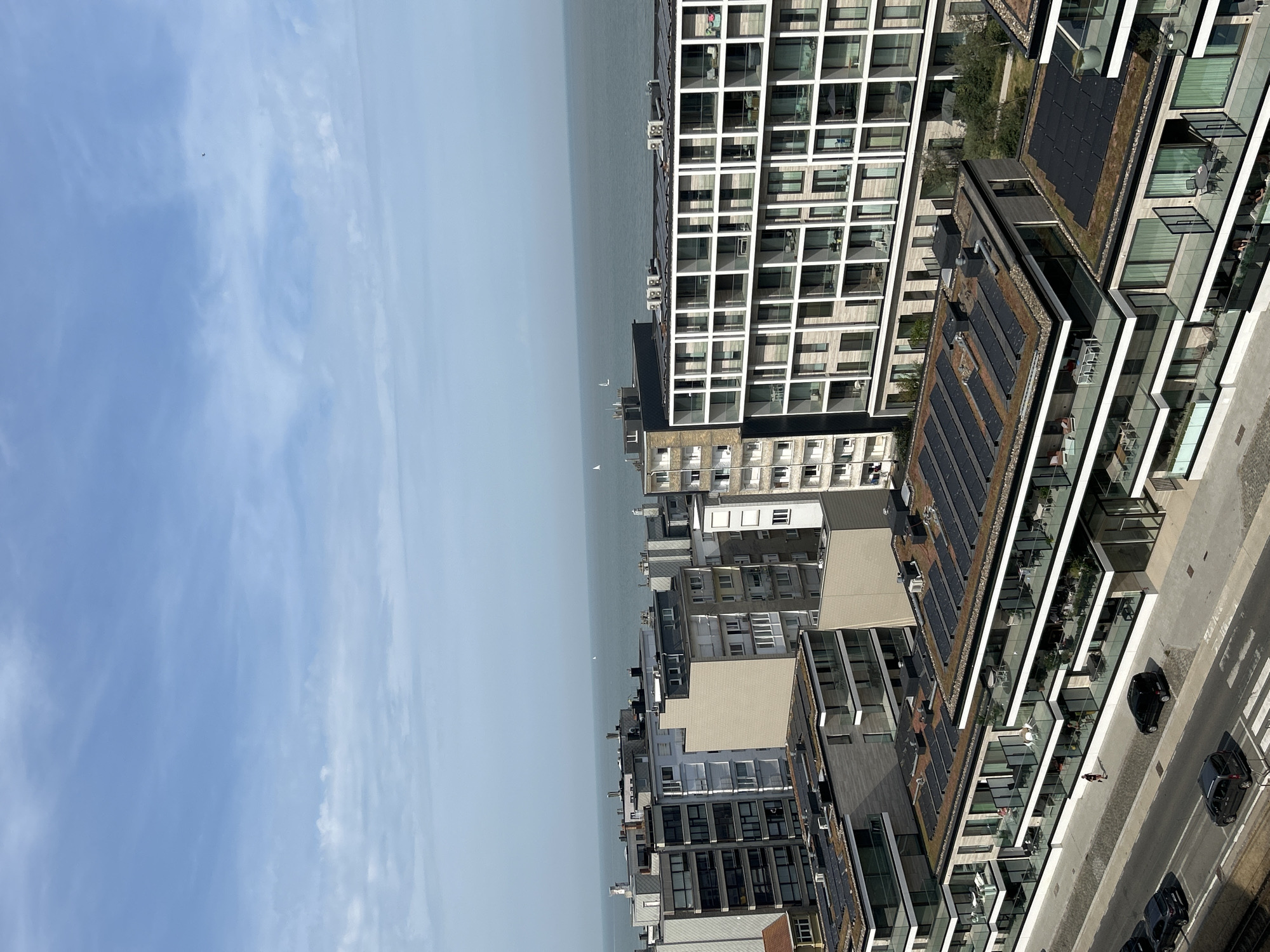 This aerial image shows a view of the coastline in Ostend, Belgium, facing the North Sea. In the foreground, a row of modern apartment buildings with flat, green roofs topped with solar panels lines a street. These buildings feature multiple stories with balconies. A few cars are parked on the street and a pedestrian walks along the sidewalk. Further back, more multi-story buildings, some appearing older and constructed with brick, are visible, also facing the sea. The sea itself is a calm, muted blue, with a few sailboats visible in the distance. The sky is a pale blue with scattered, wispy white clouds.