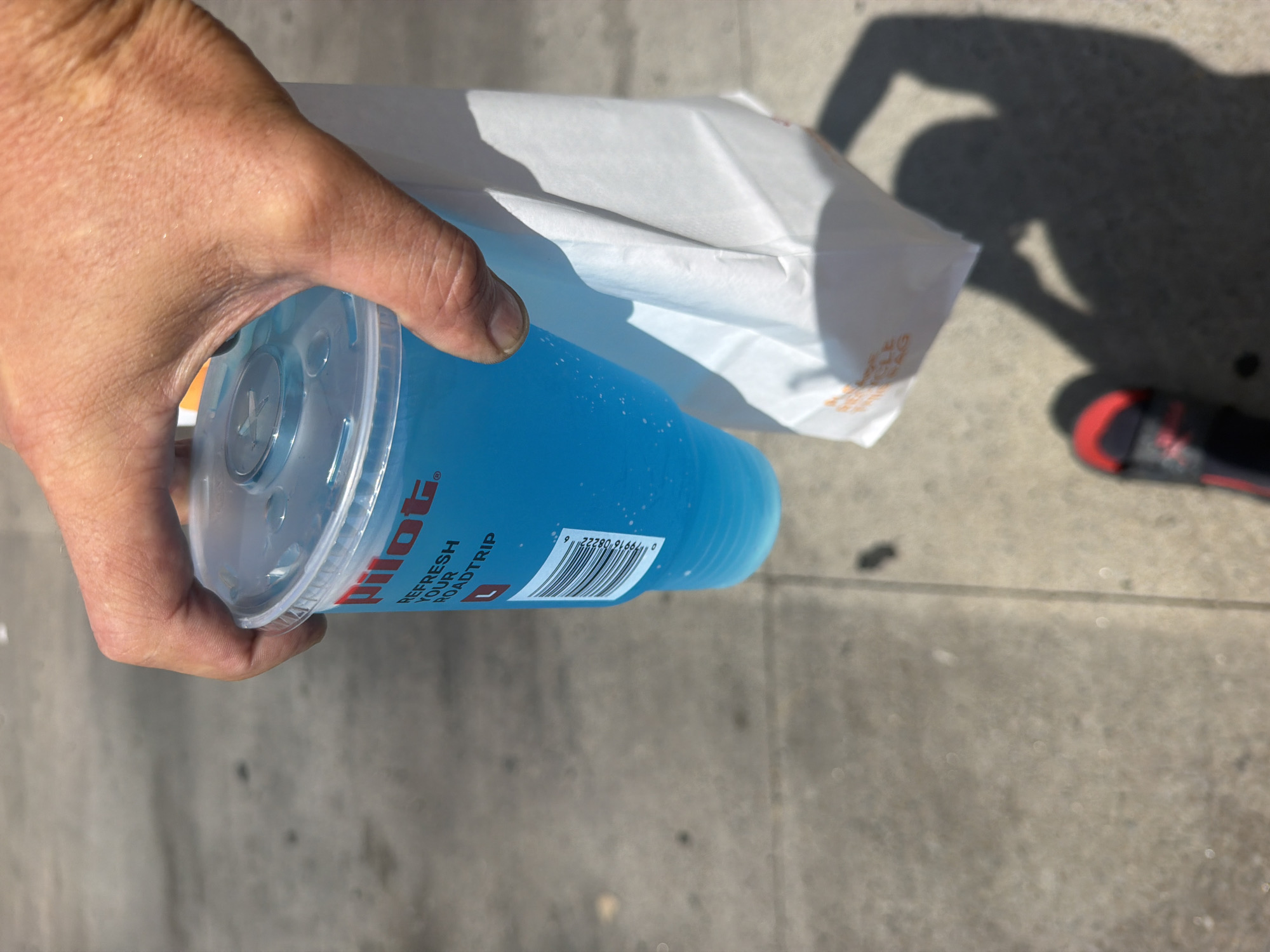 A person's hand is holding a large, clear plastic cup filled with a bright blue beverage. The cup has a clear plastic lid with a circular opening. The logo "Pilot" is visible in red on the side of the cup, along with text that reads "REFRESH YOUR ROADTRIP." A white paper bag with orange lettering is tucked under the cup, held by the same hand. The background is a paved surface, likely a sidewalk or road, with shadows cast by the sun. A shoe with a black band and red accents is visible in the lower part of the frame. The scene appears to be outdoors during the day. The location is the city of Hayti, United States of America.